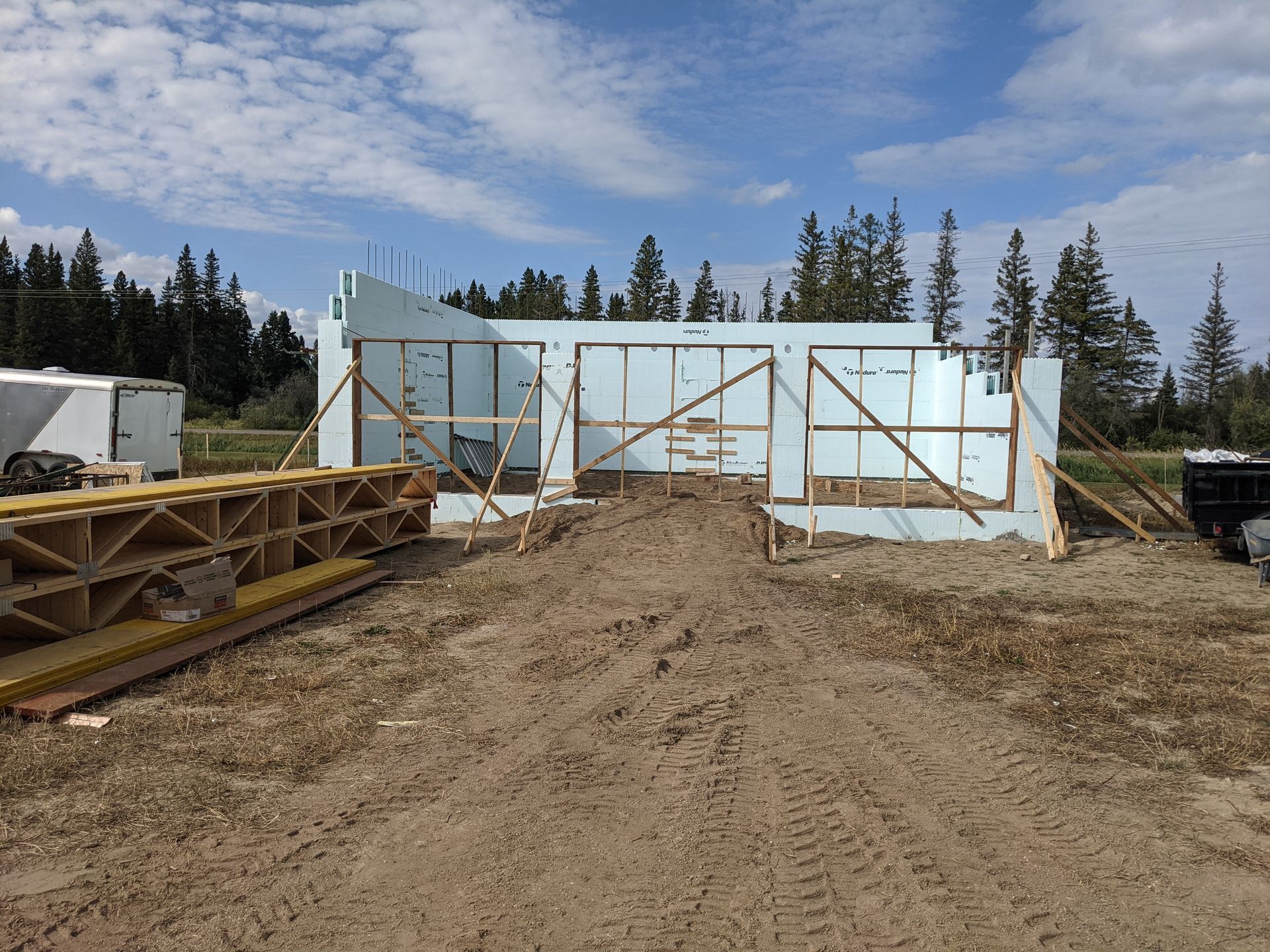 Construction site with insulated concrete forms, dirt pile, wooden supports, and trees in background.