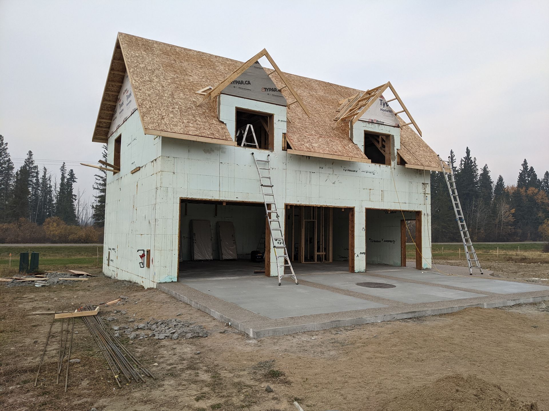Two-story garage under construction; exterior walls covered in white insulation, unfinished roof, concrete pad.