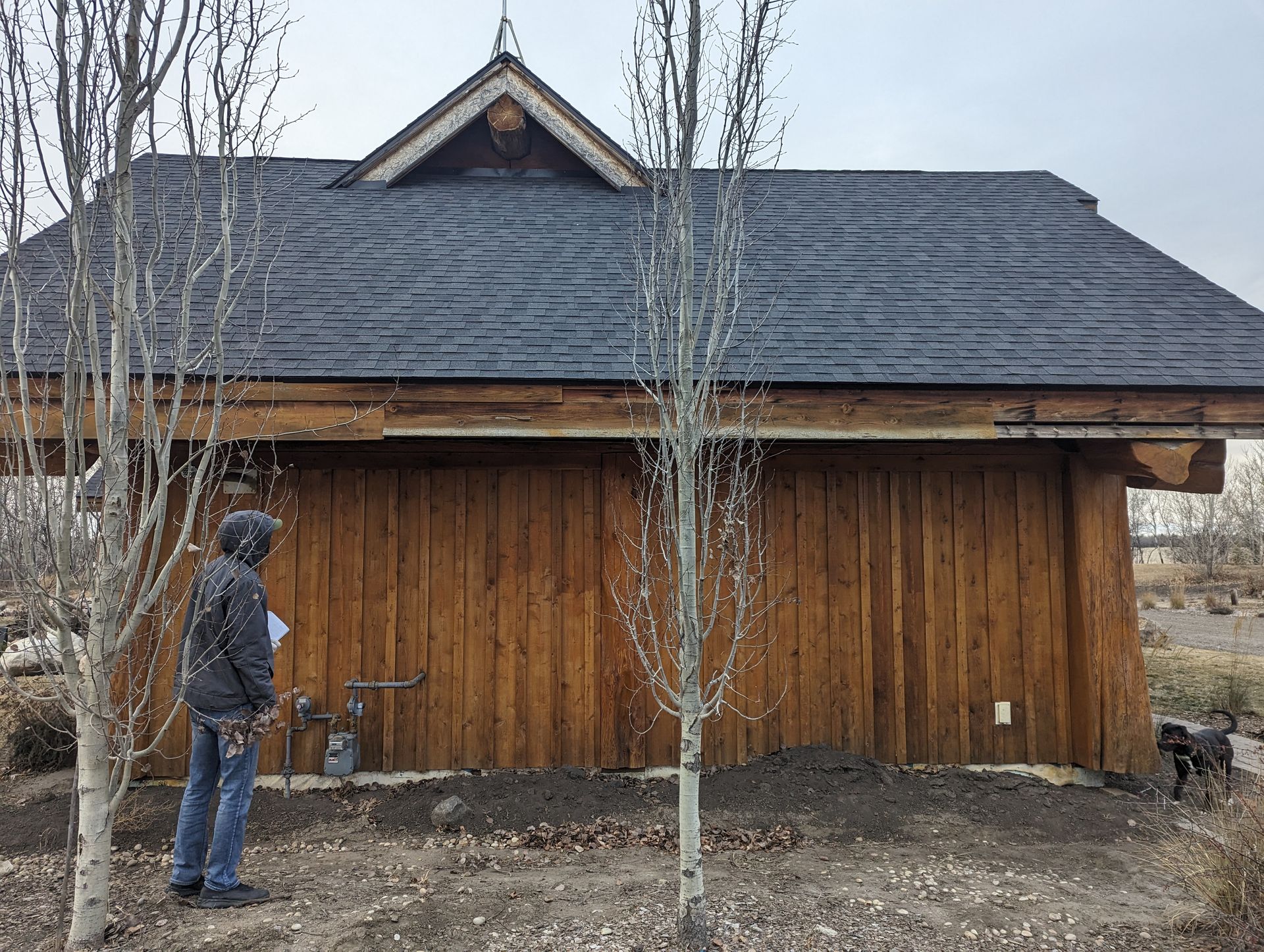 Man near wooden building with black roof; trees in front.
