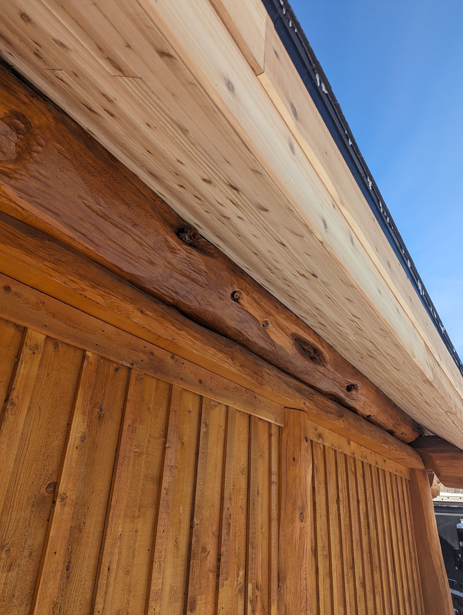 Wooden cabin exterior with log beams and plank ceiling against a blue sky.