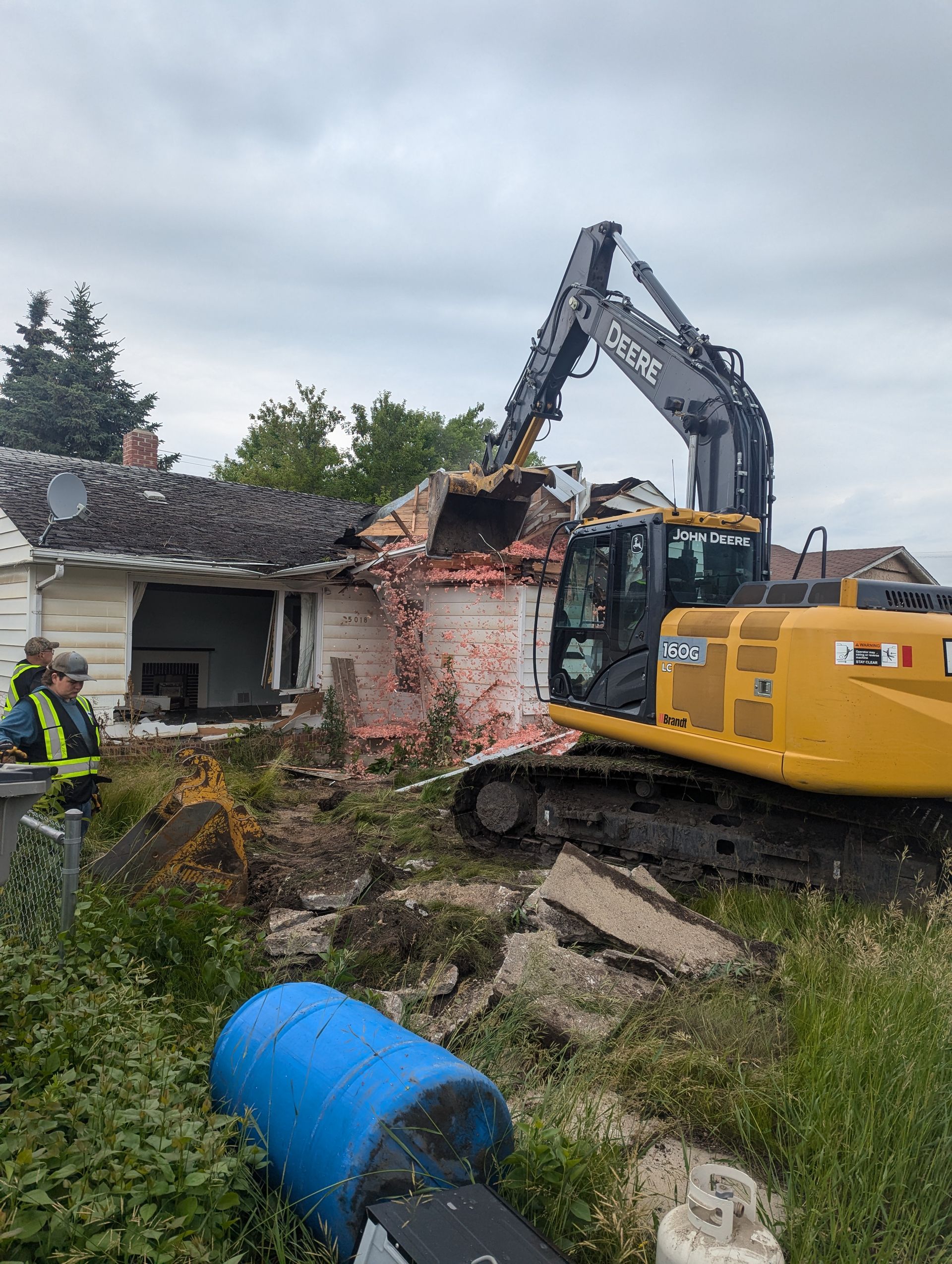 Construction excavator demolishing a small brick house; two workers in vests watch. Overcast sky.