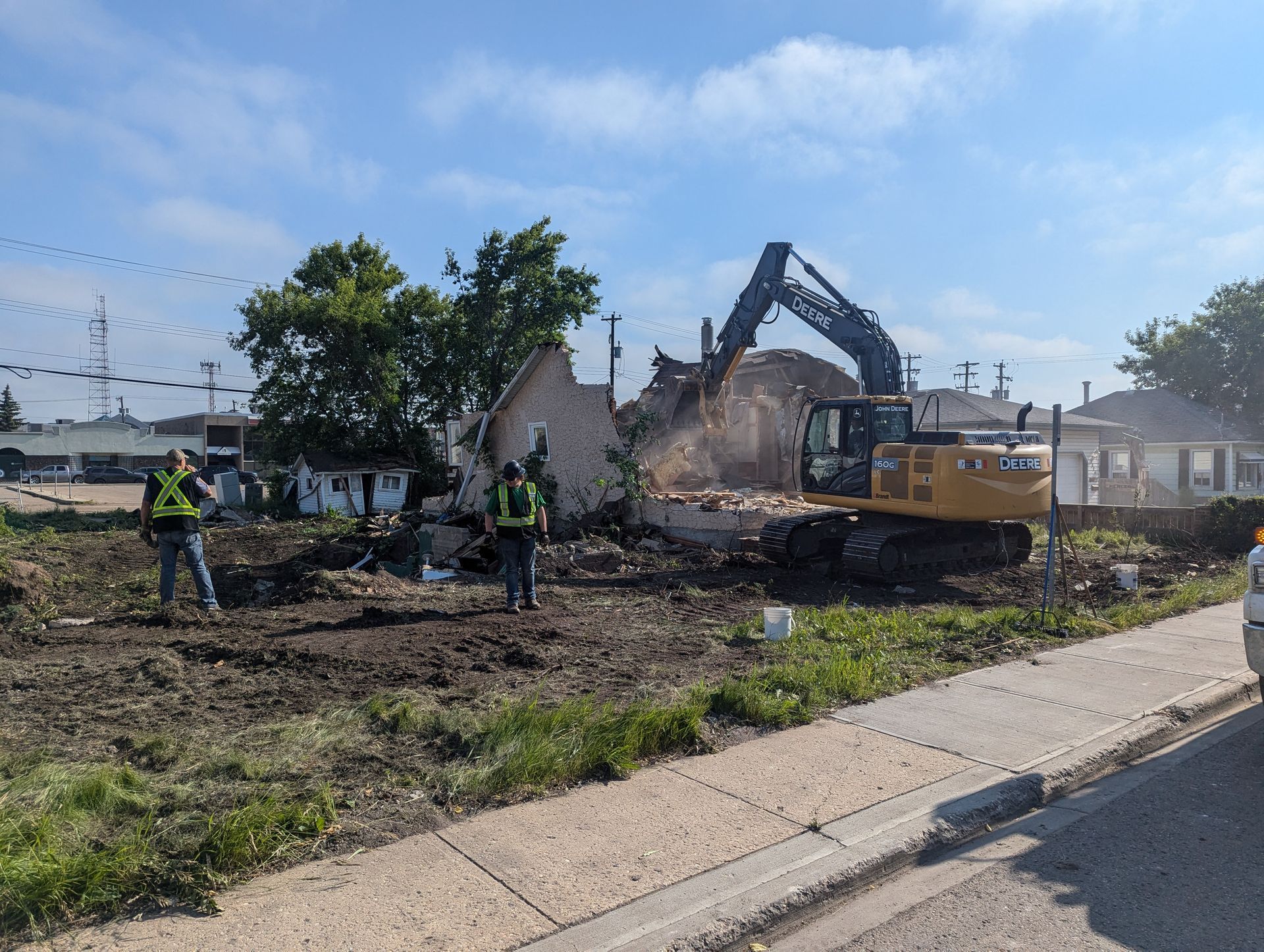 Excavator demolishes a building; two workers in vests watch from the dirt lot. Clear sky.