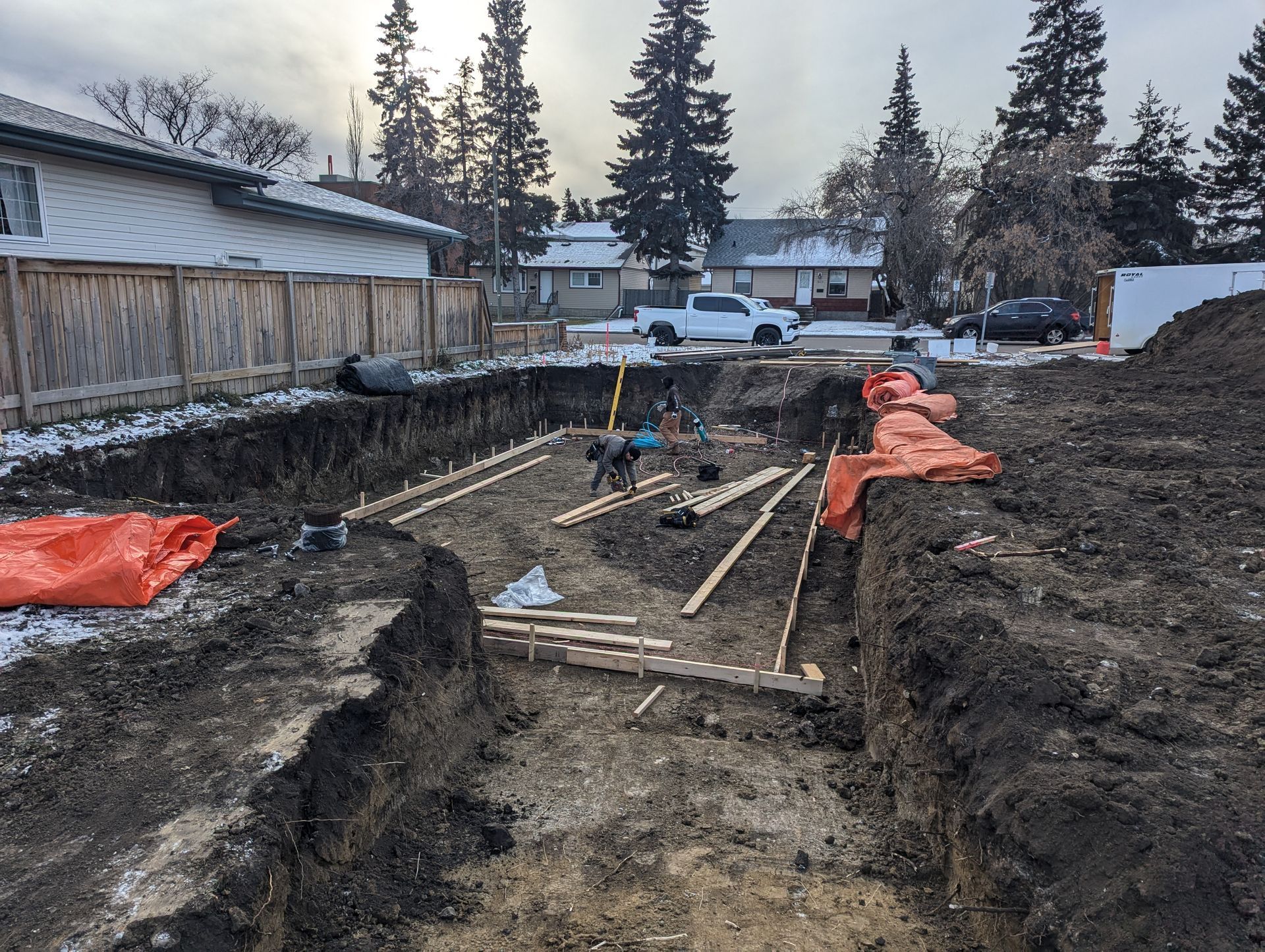 Construction site, foundation excavation in residential area. Trenches, lumber, tools, orange safety coverings, and soil.