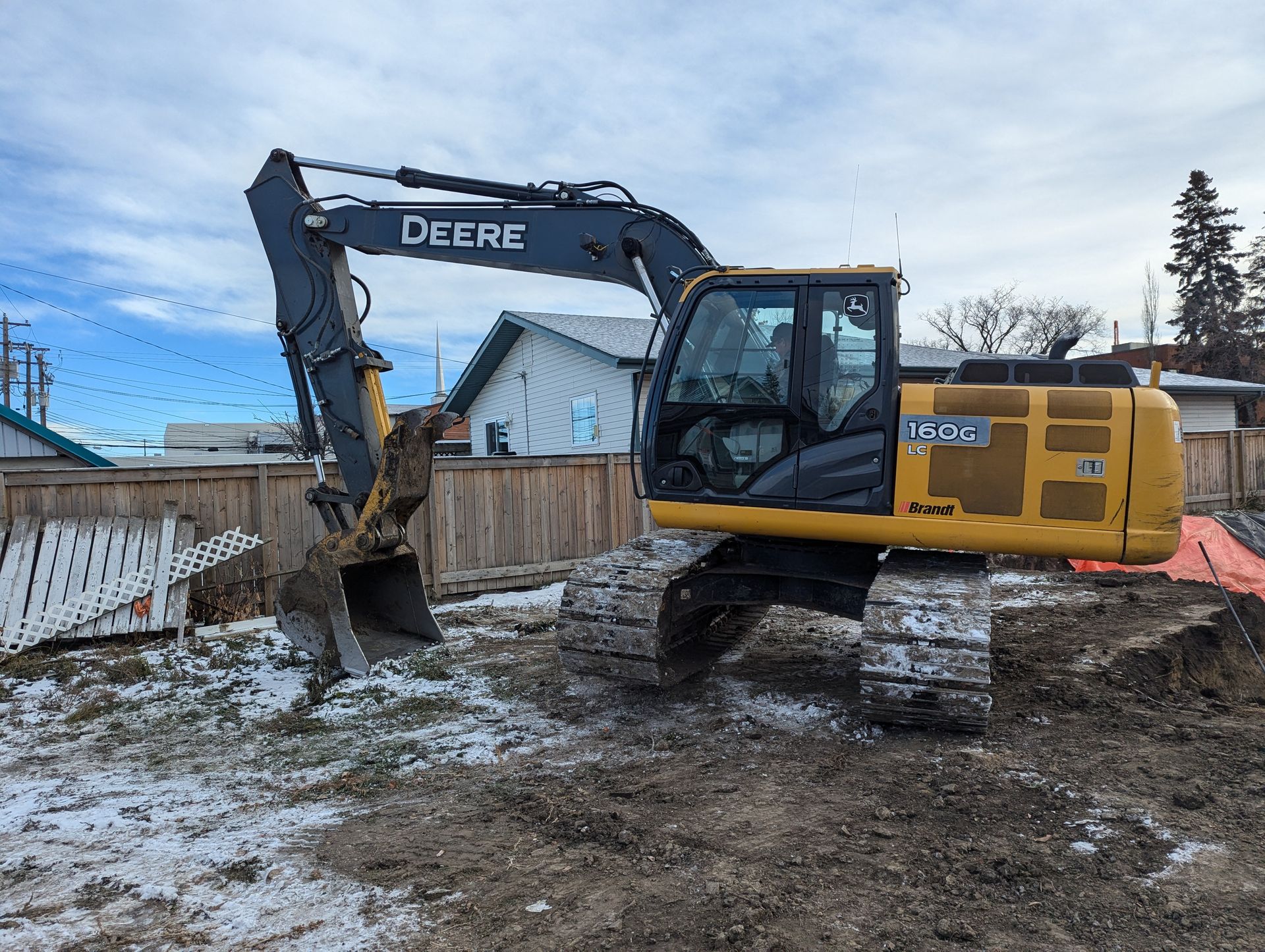 Yellow and black John Deere excavator at a construction site with wooden fence and a blue sky in the background.