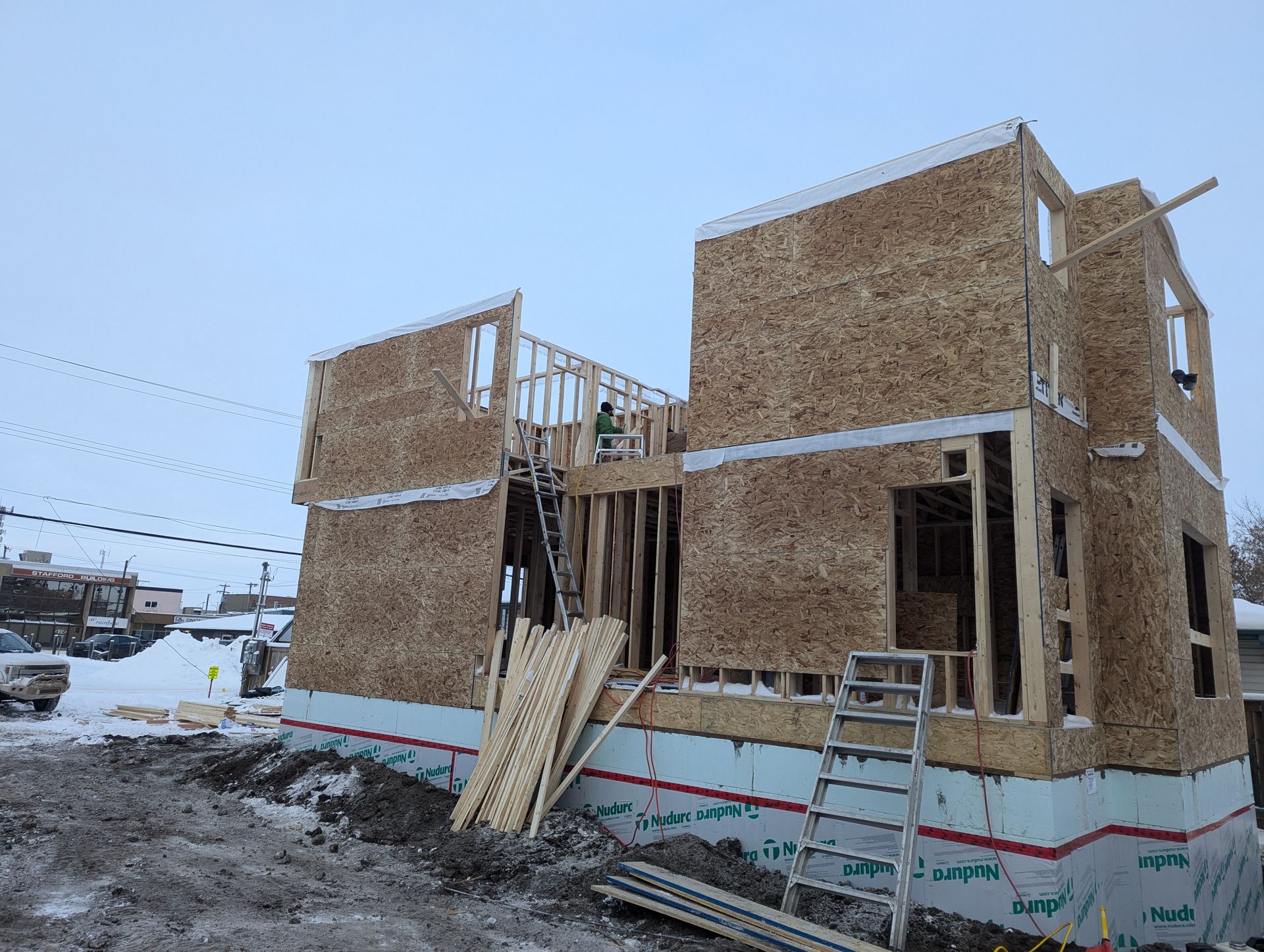 Construction of a two-story building in progress, framed with plywood and wood studs. Exterior insulation and snow present.