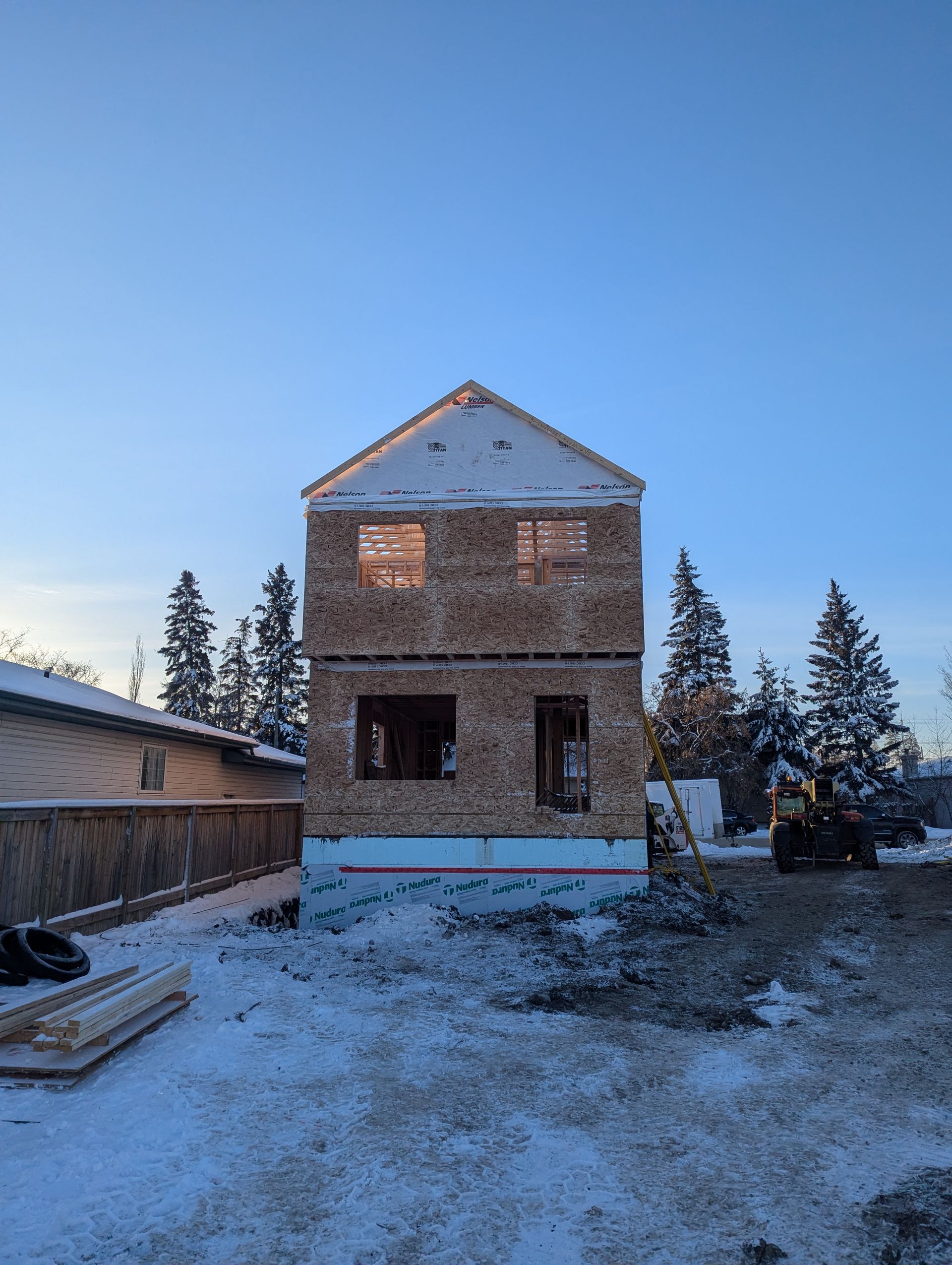 Two-story house under construction in winter; snow-covered ground, blue sky, wooden frame.