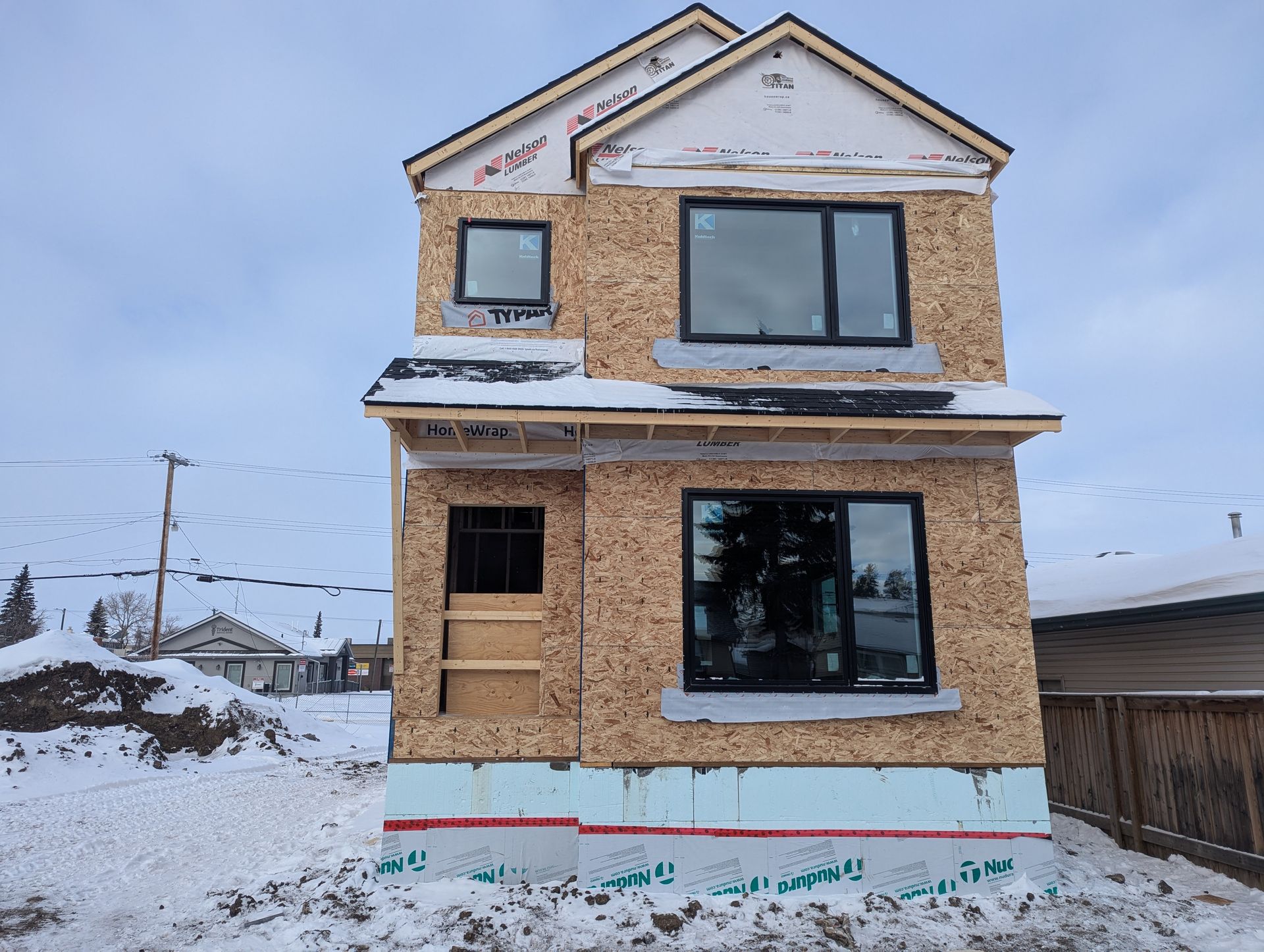 Two-story house under construction with exposed wood panels, black windows, and snow on the ground.