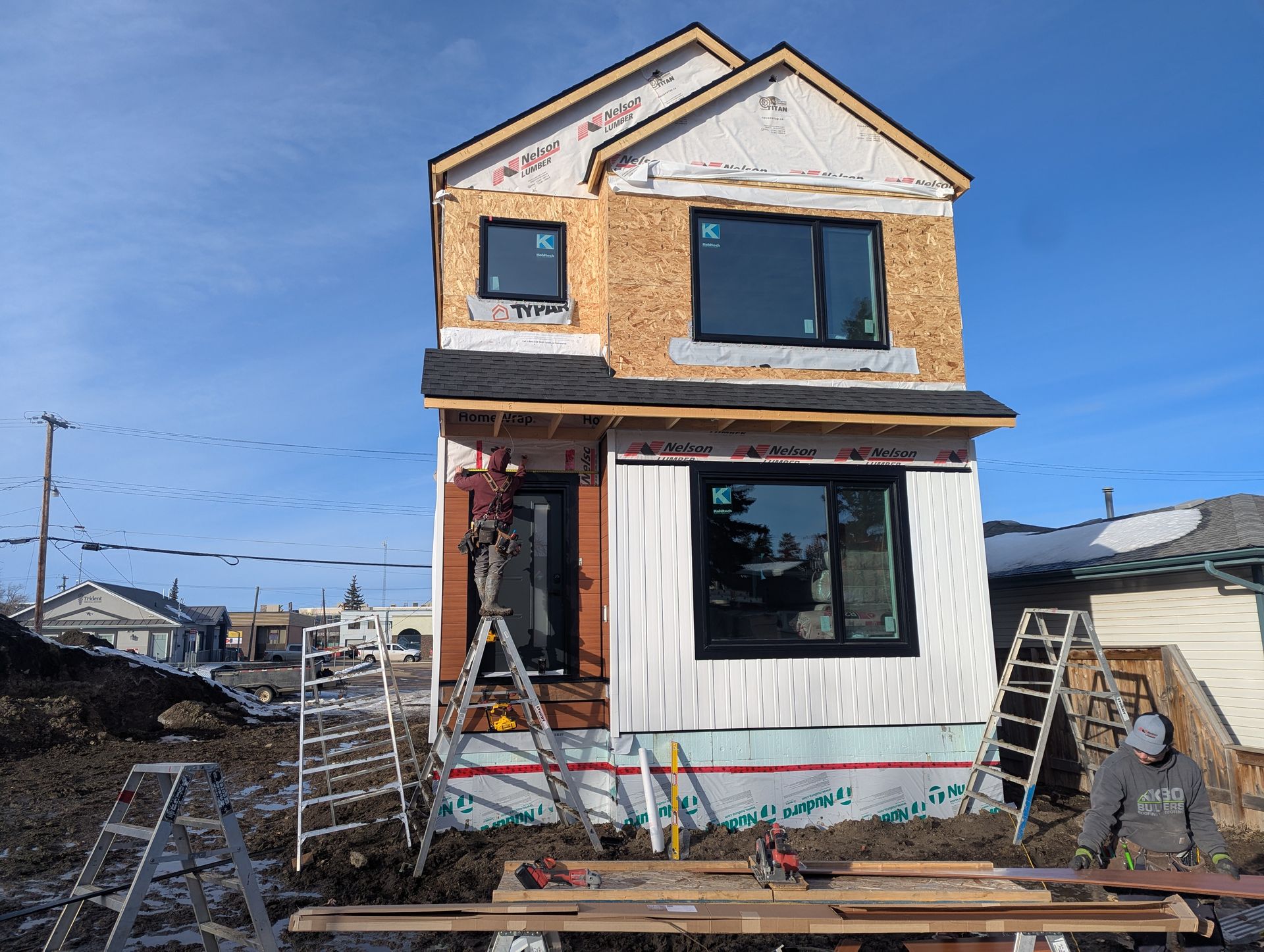 Two-story house under construction, with exposed wood framing, black windows, and blue sky.