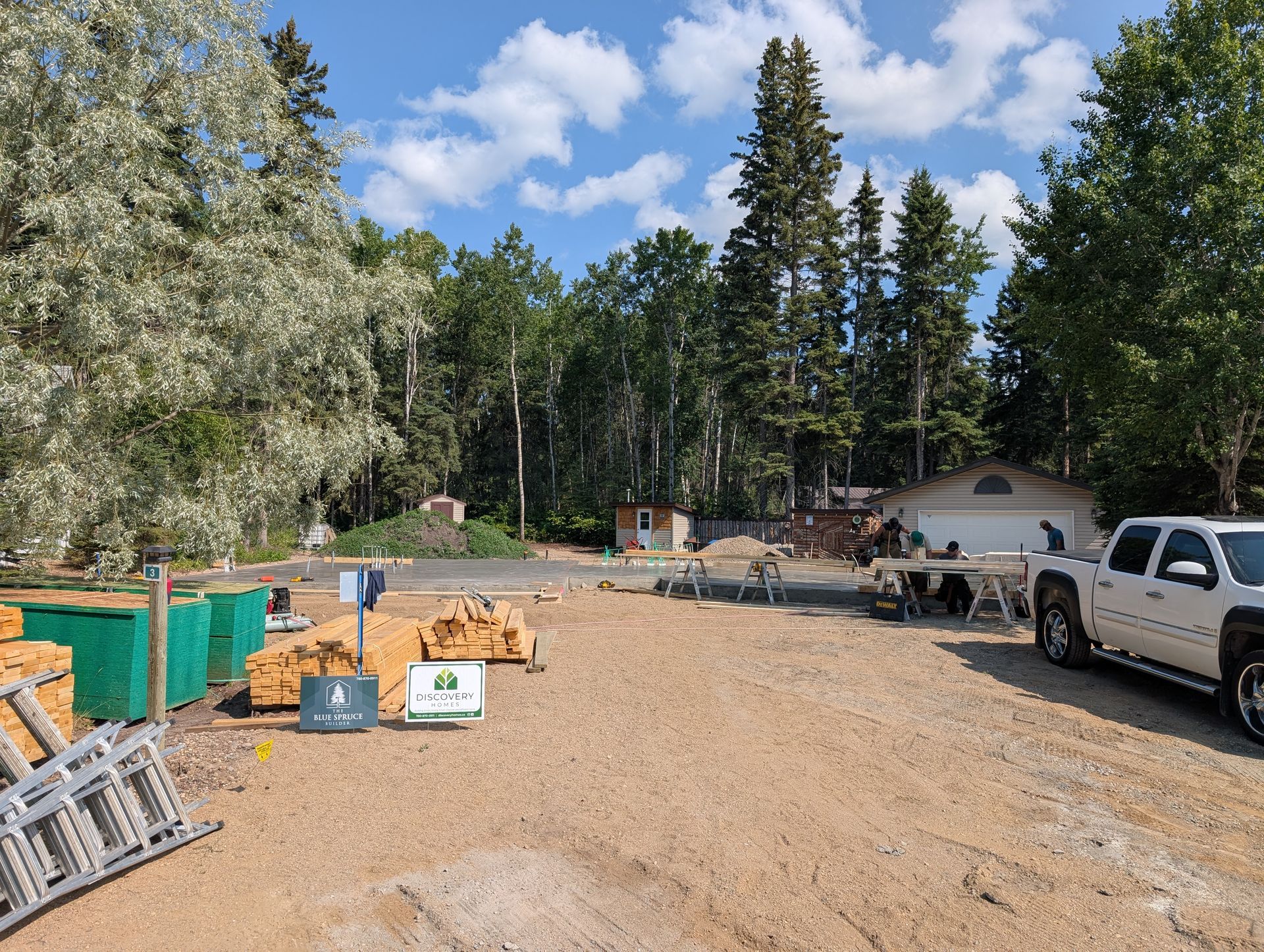 Gravel lot with lumber, equipment, and a white pickup truck; trees and sky in the background.