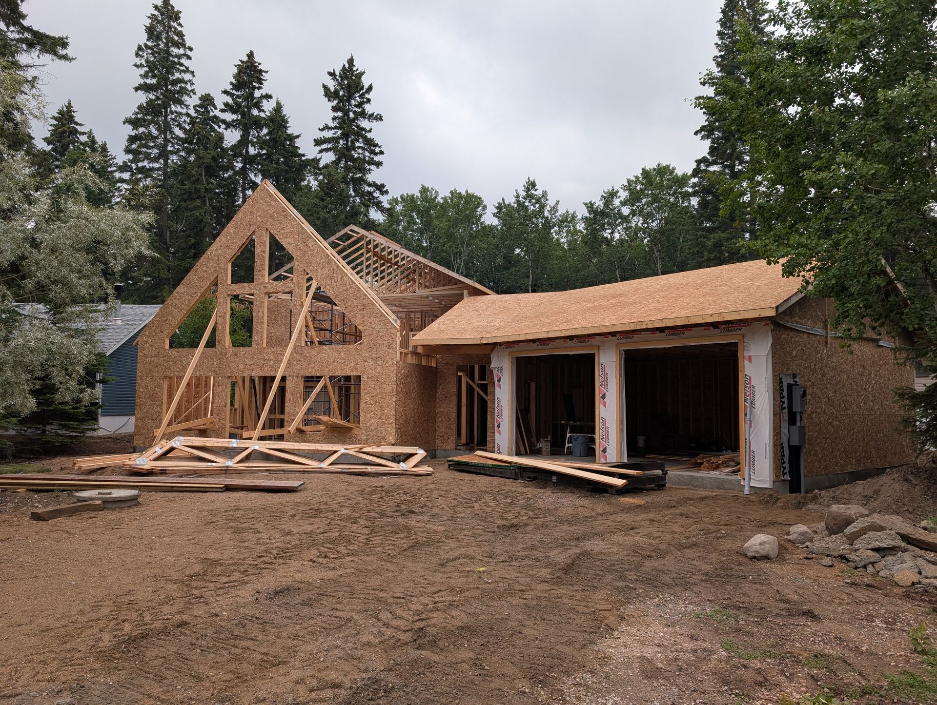House under construction, wood frame visible. Garage and main structure with open roof. Brown ground, trees in the background.