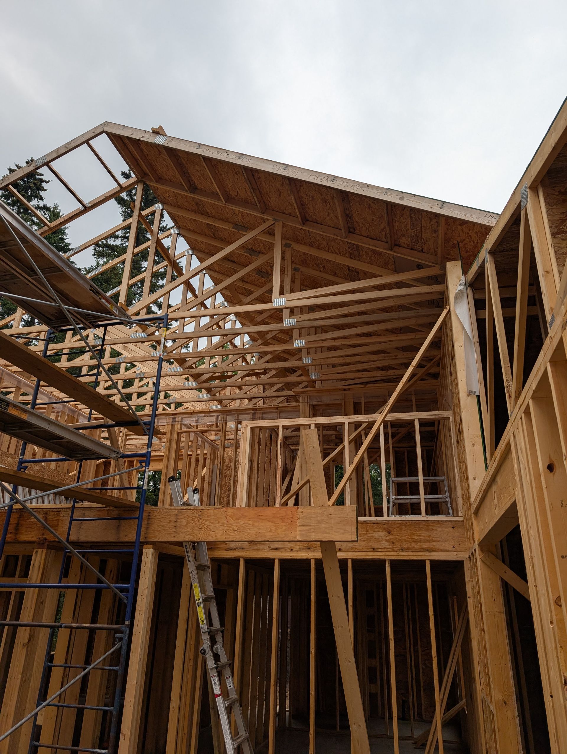 Wooden house frame under construction, with exposed rafters and scaffolding against a cloudy sky.