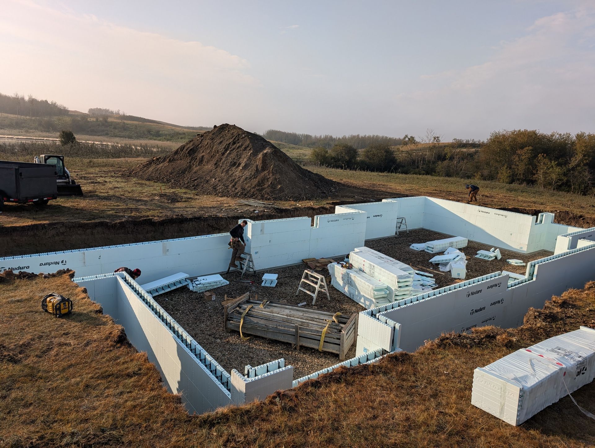 Construction site with white foam block foundation walls. Workers inside, dirt pile in background.