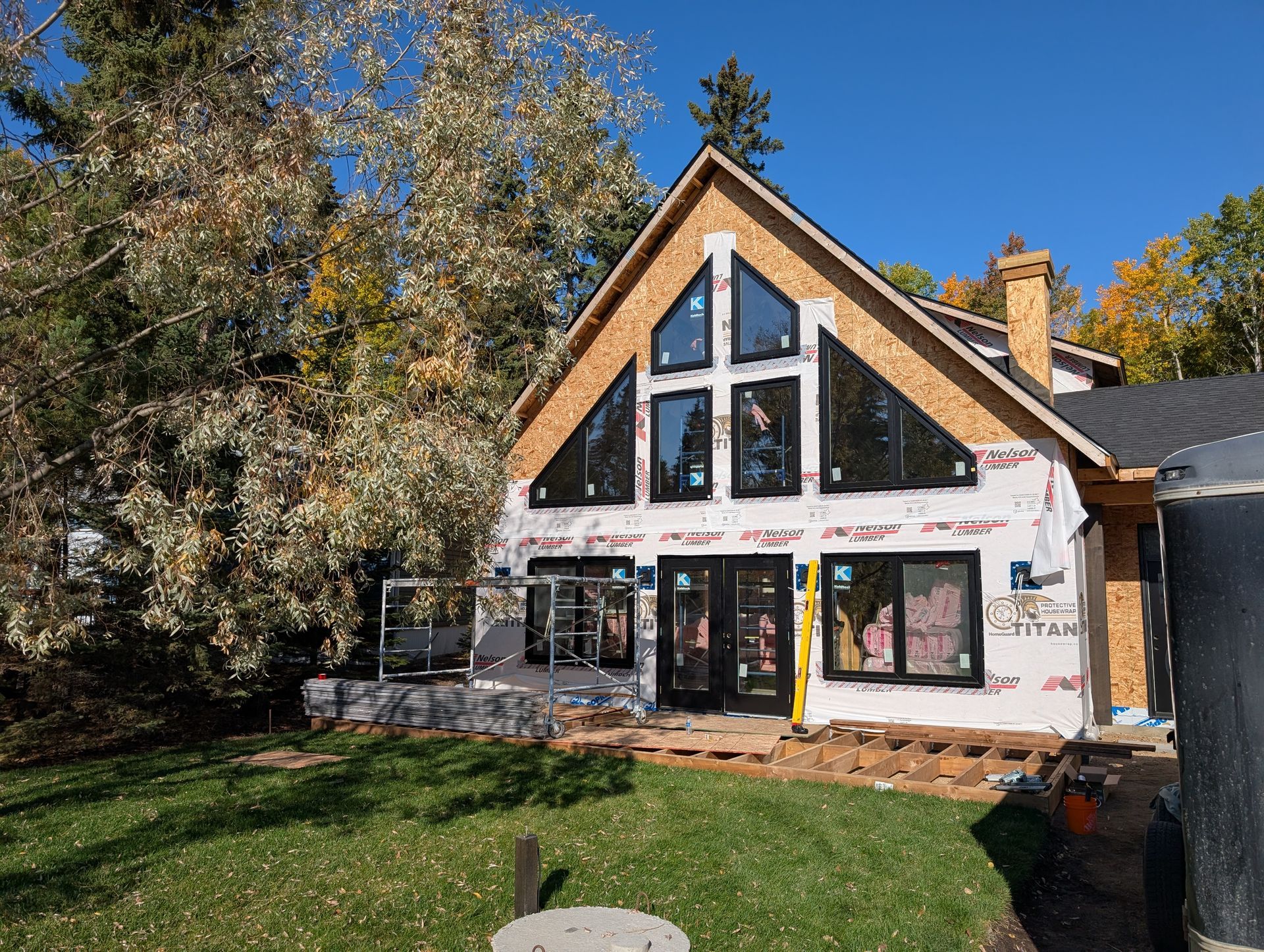 House under construction with large windows and a gable roof, surrounded by trees and green grass.