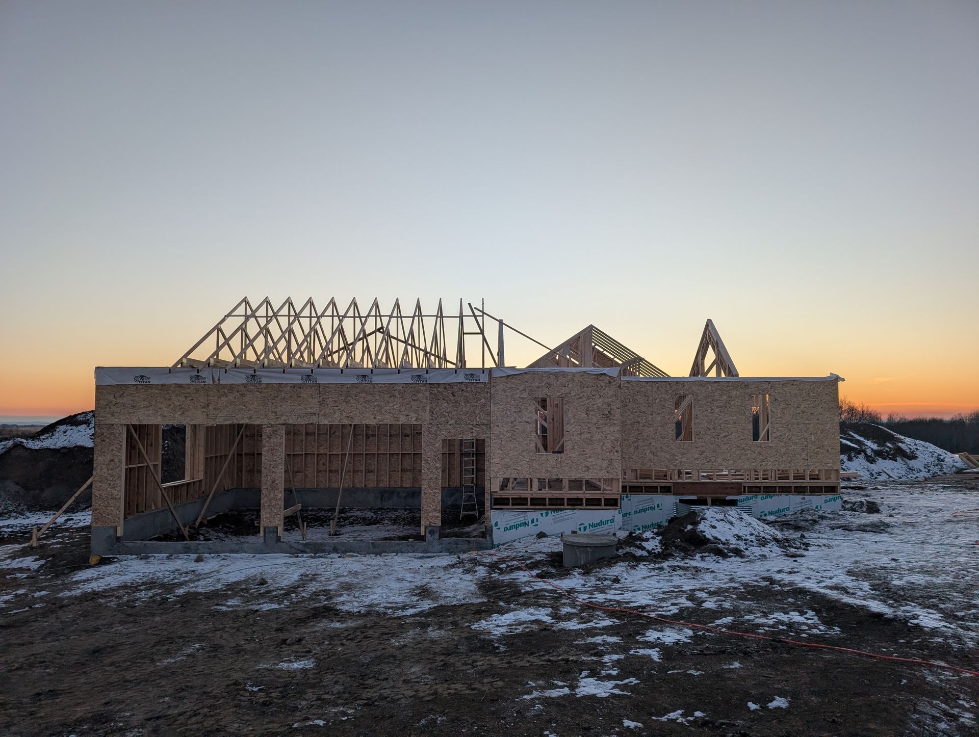 New house under construction with exposed wooden frame, in snowy field at sunset.