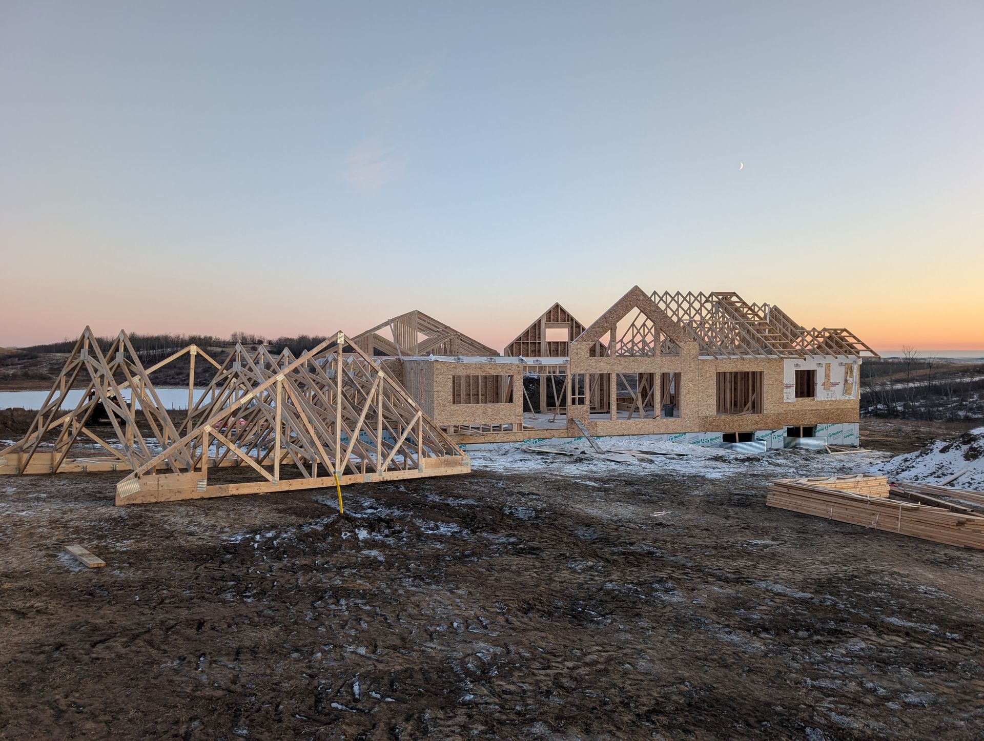 House under construction; wooden frame and trusses, snowy ground, dusk sky.