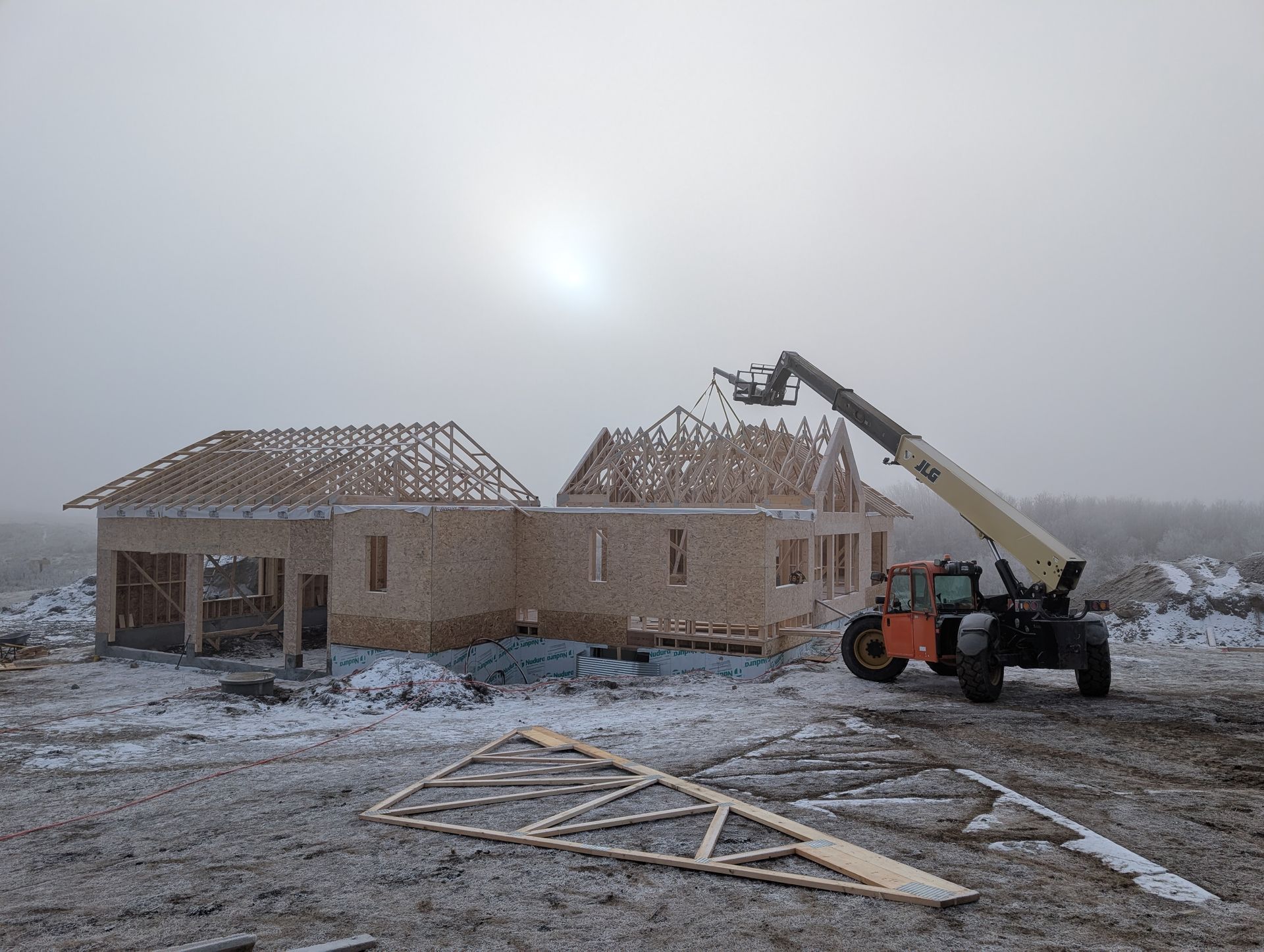 House under construction on a snowy day; a telescopic handler places roof trusses.