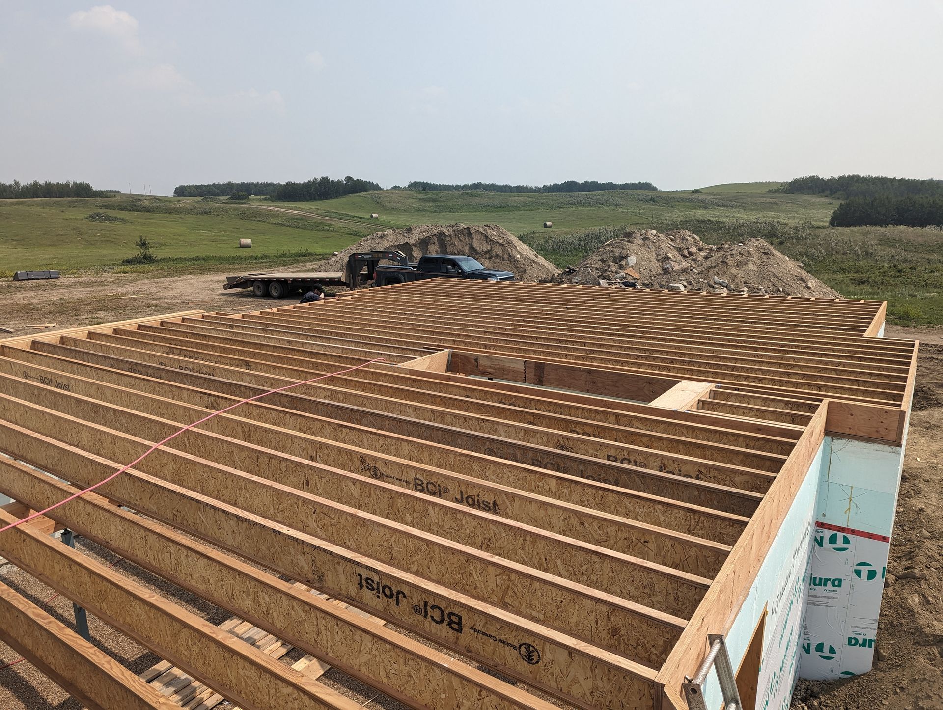 Wooden house foundation under construction with lumber, sky, and rural landscape.