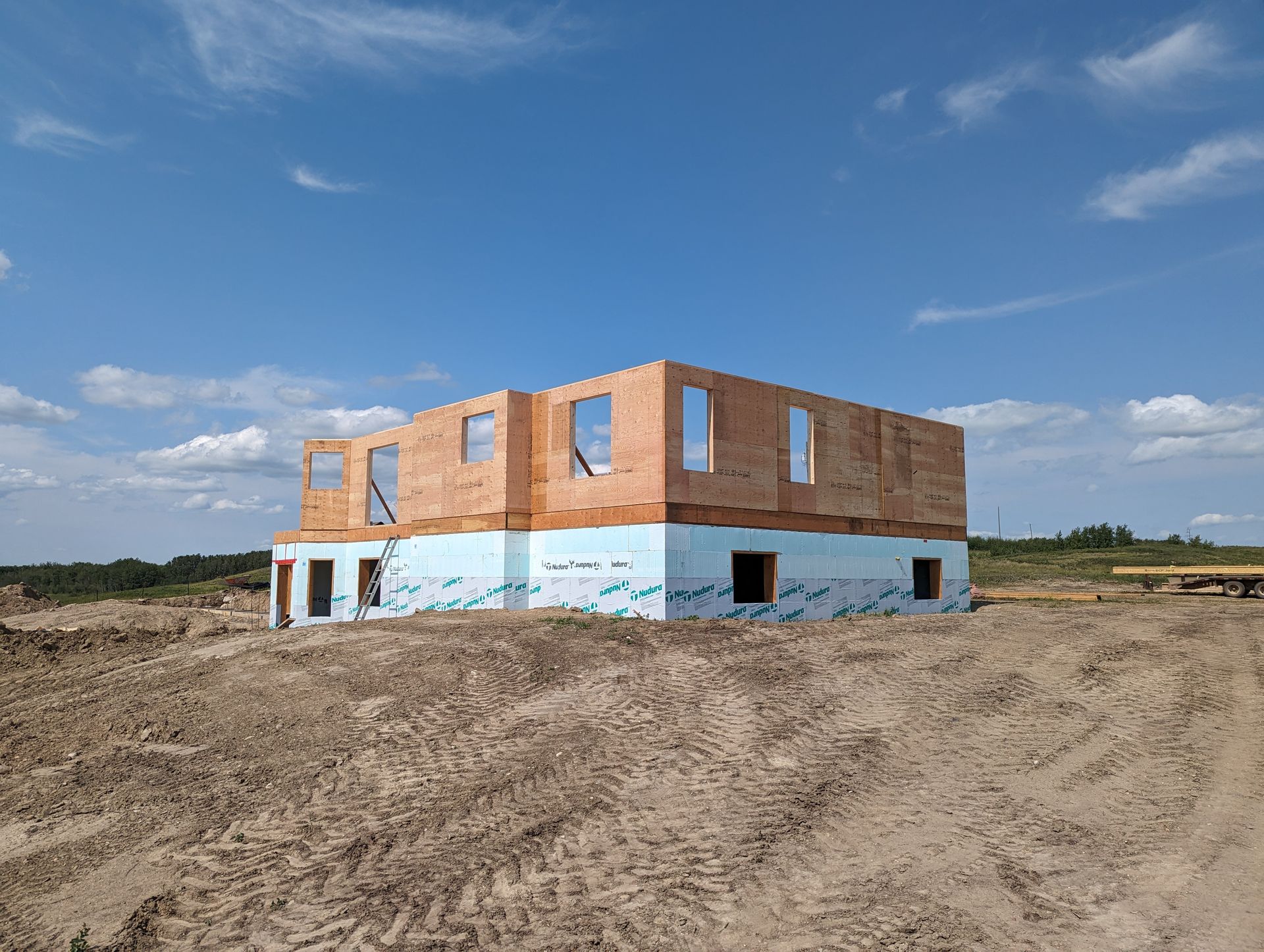 Two-story house under construction on a dirt lot, blue insulation on the first floor, brickwork on top, and blue sky.