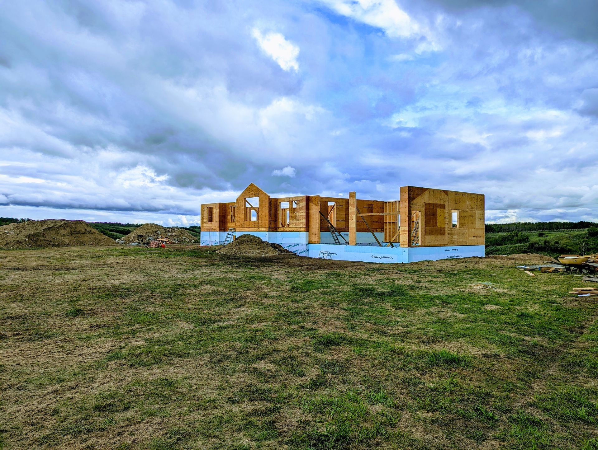 Construction of a wooden house frame on a grassy plain under a cloudy sky.