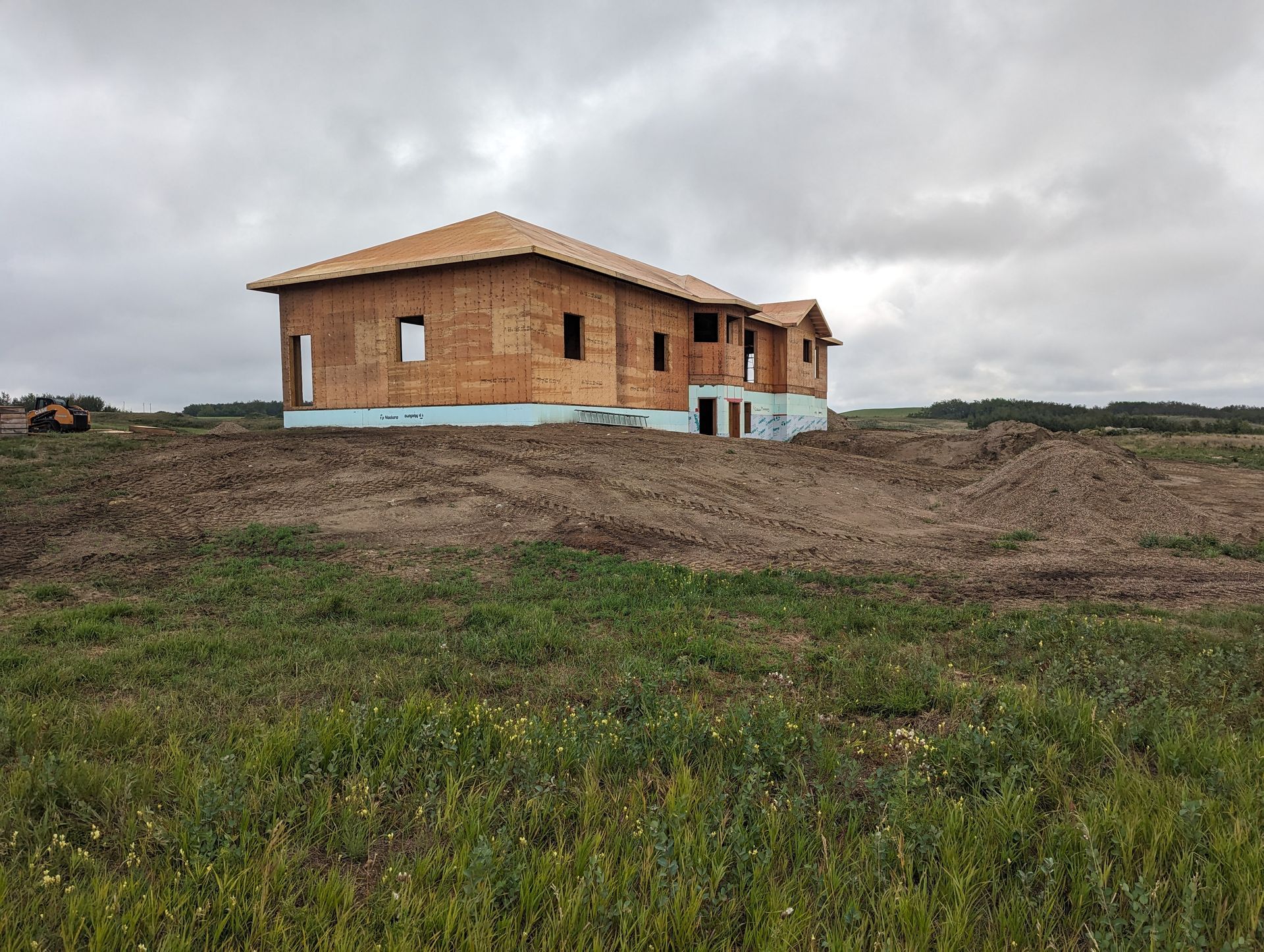 Unfinished house under construction on a grassy hill, against a cloudy sky.