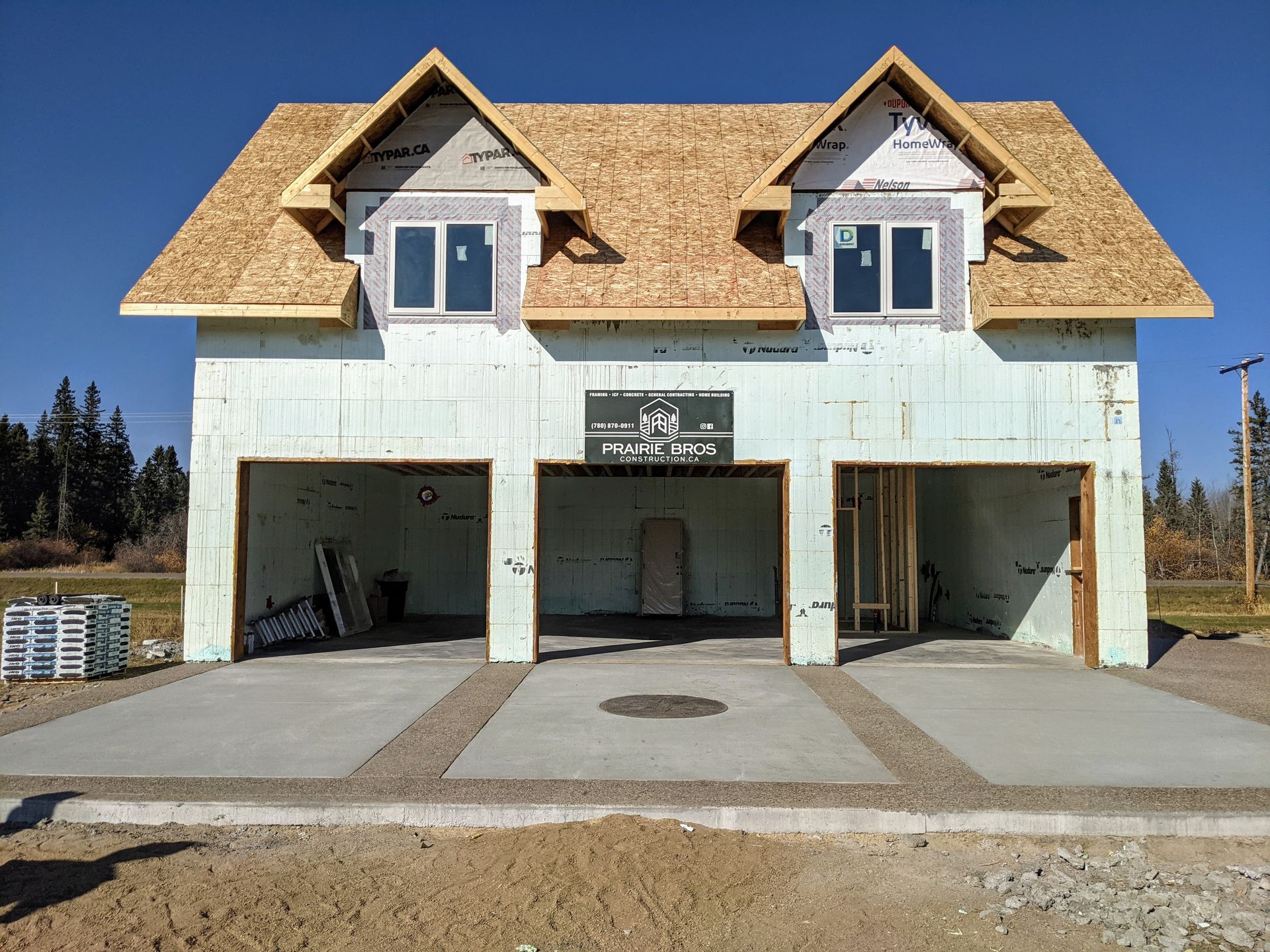 Under construction two-story garage with three bays and two dormers. Blue insulation, concrete driveway, and brown roof.