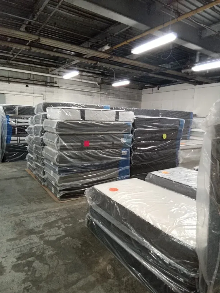 Stacked plastic-wrapped mattresses in a warehouse under bright overhead lights.