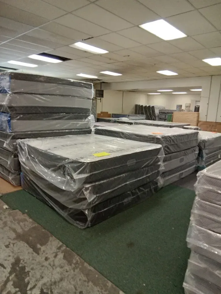 Stacked mattresses wrapped in plastic inside a warehouse with green carpet and fluorescent lighting