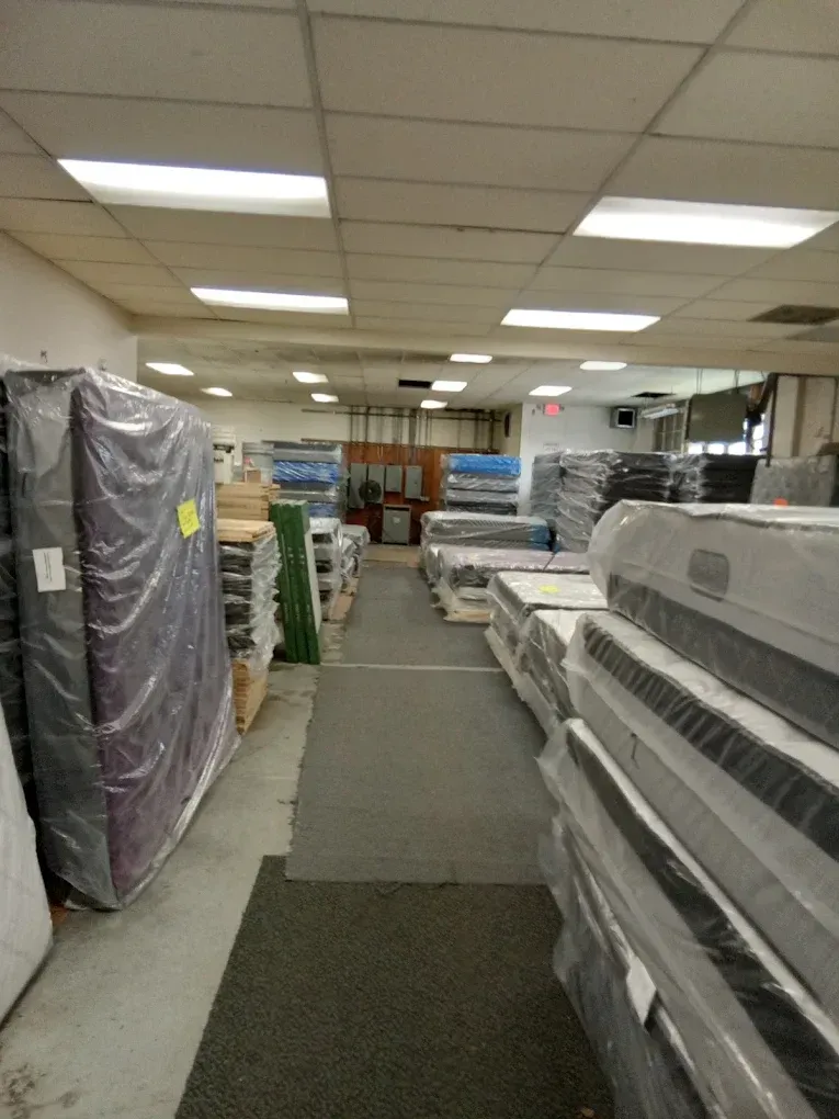 A furniture store aisle with stacked mattresses under fluorescent ceiling lights.