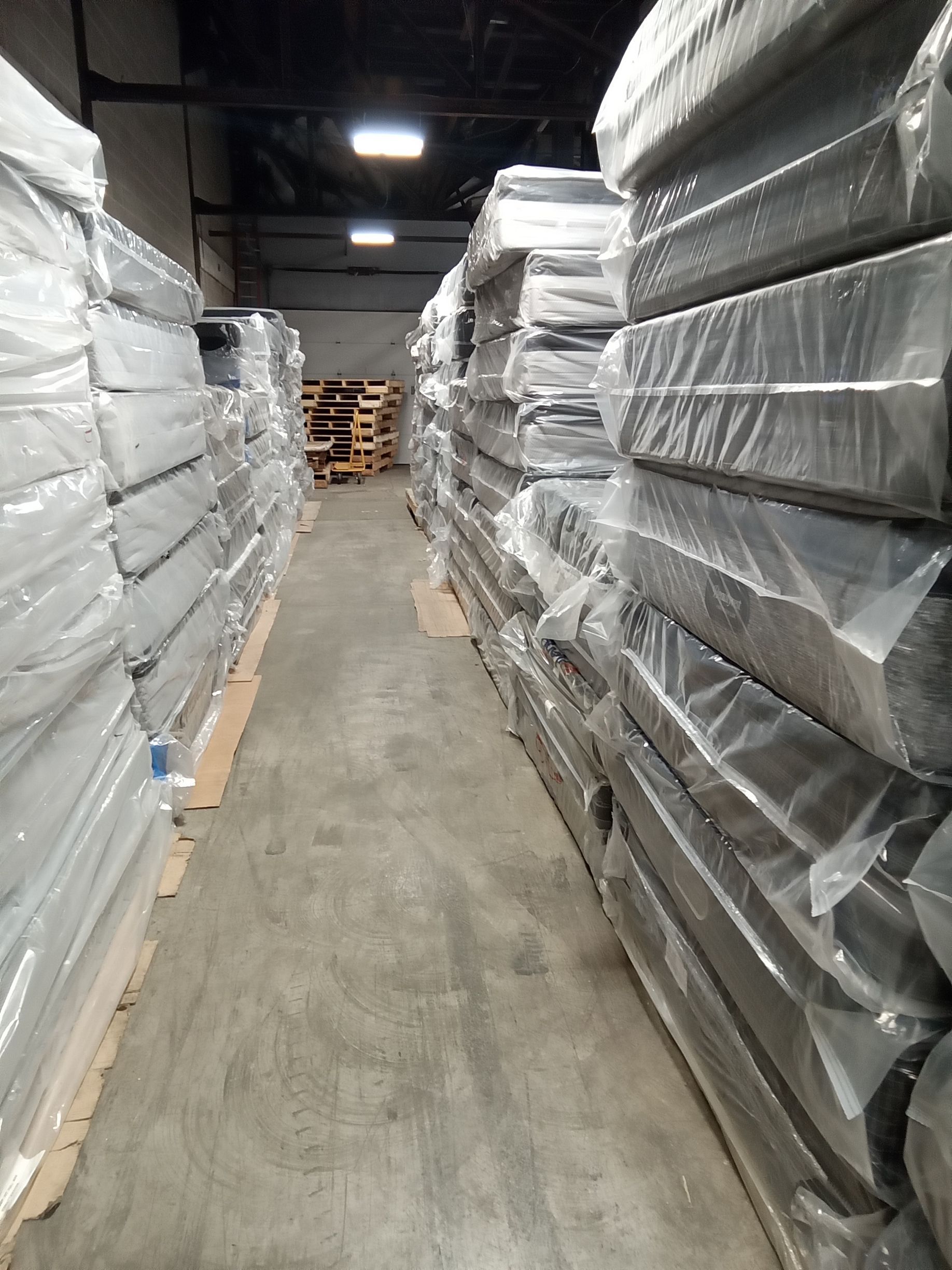 Narrow warehouse aisle lined with stacked, plastic-wrapped pallets and boxes on both sides.