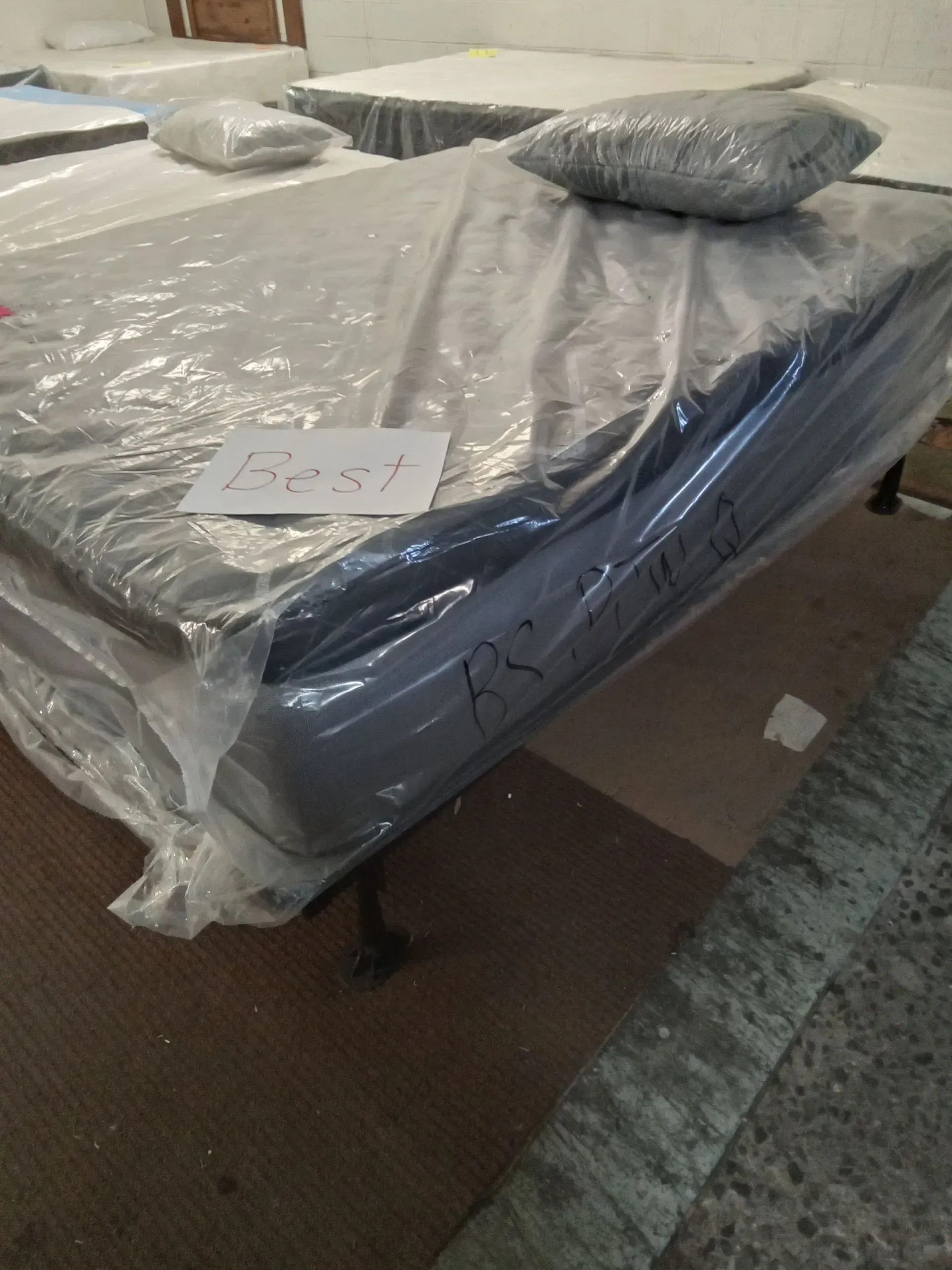 Wrapped mattress on a bed frame with a dark pillow in a room with carpeted floor