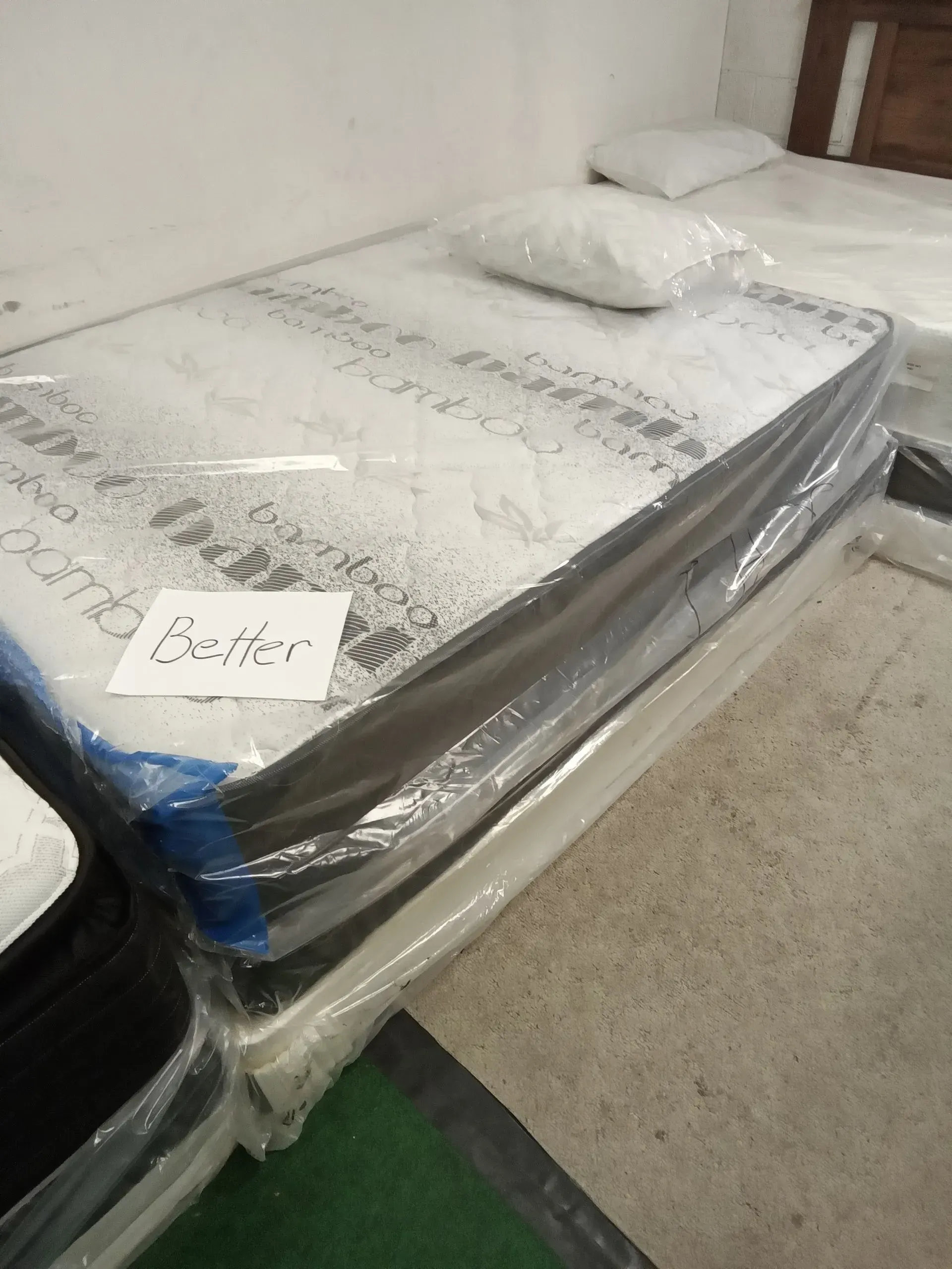 Plastic-wrapped mattress on a floor, with two pillows on top and a small label attached