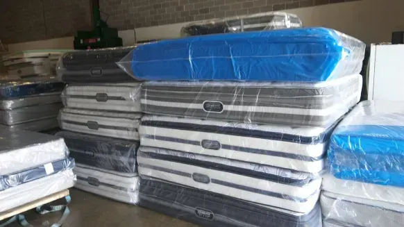 Stacked mattresses wrapped in plastic, mostly white with blue covers, stored indoors on shelves.