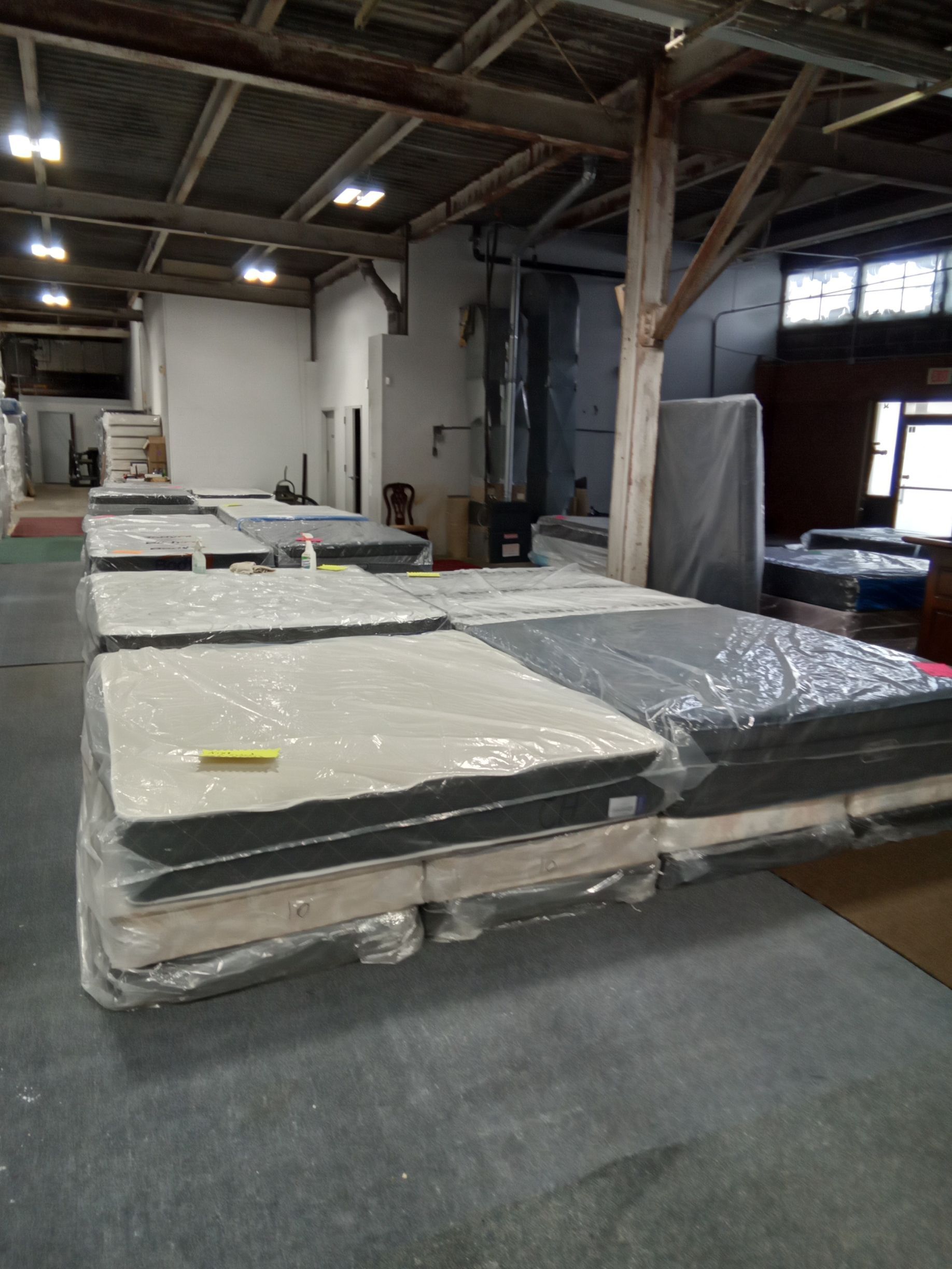 Warehouse with stacked plastic-wrapped mattresses or foam panels on pallets under industrial lighting