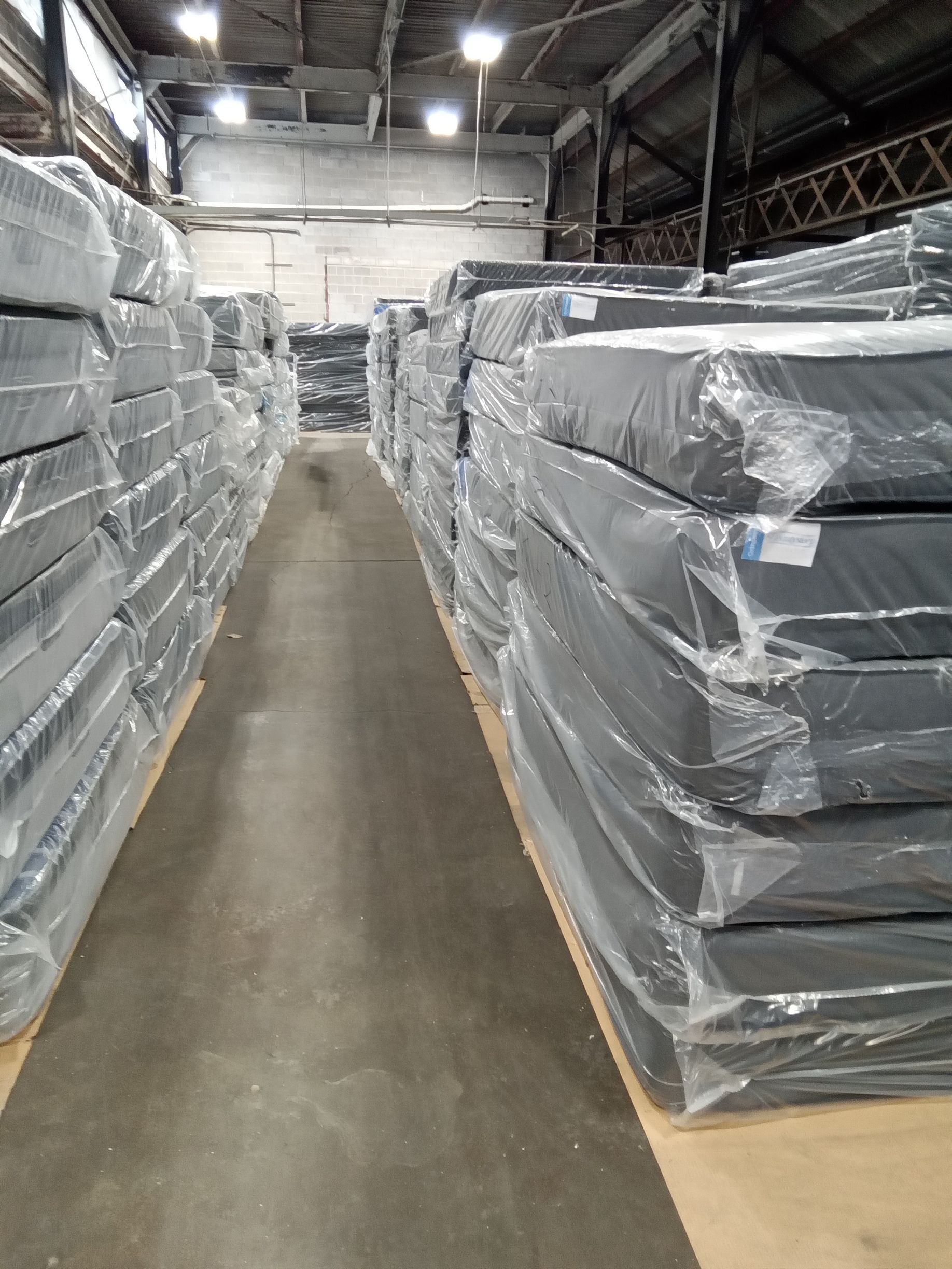 Warehouse aisle lined with stacked pallets wrapped in gray plastic under bright overhead lights