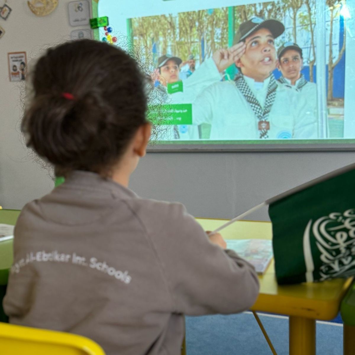 A girl wearing a jacket that says ' schools ' on the back sits at a table