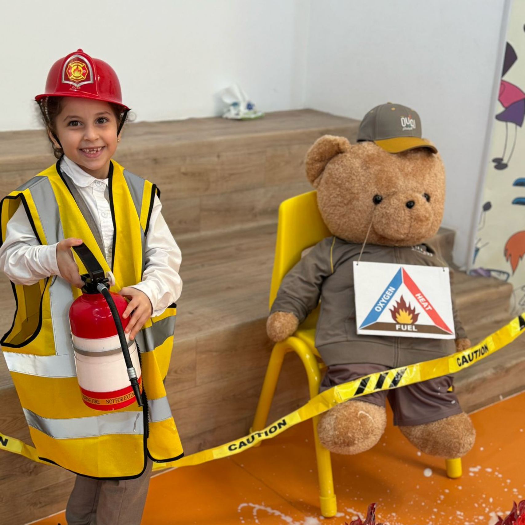 A child dressed as a fireman holding a fire extinguisher next to a teddy bear