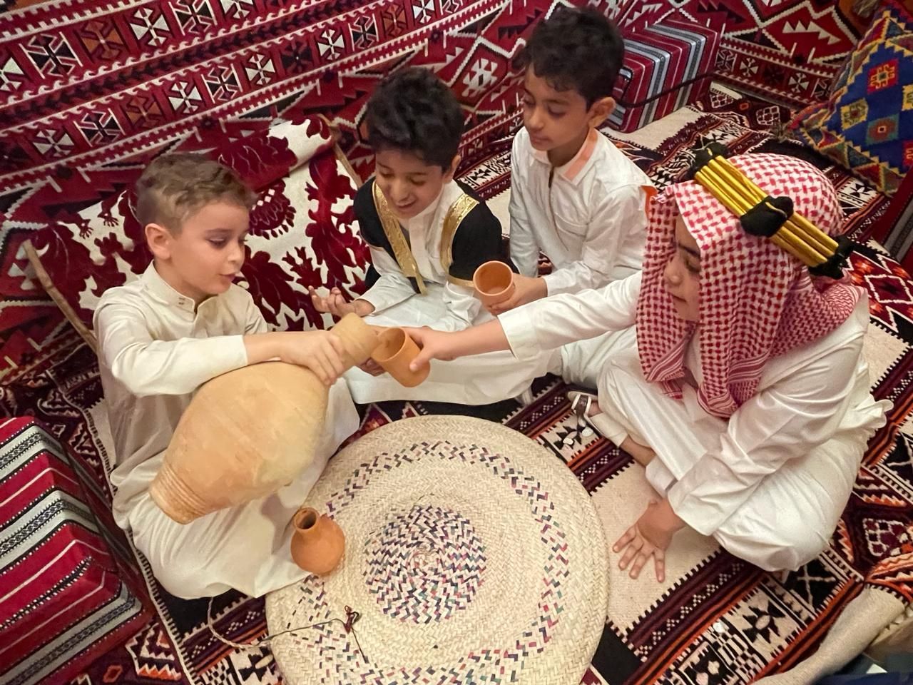 A group of children are sitting on a rug drinking from clay pots.