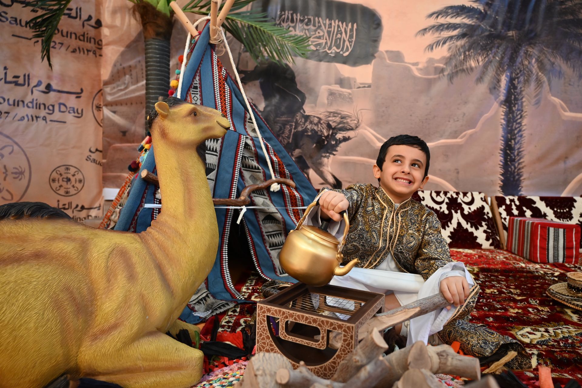 A boy is sitting in front of a camel statue holding a teapot.