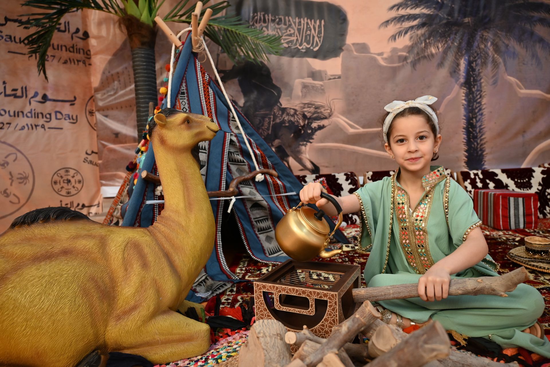 A little girl is sitting in front of a camel statue holding a teapot.