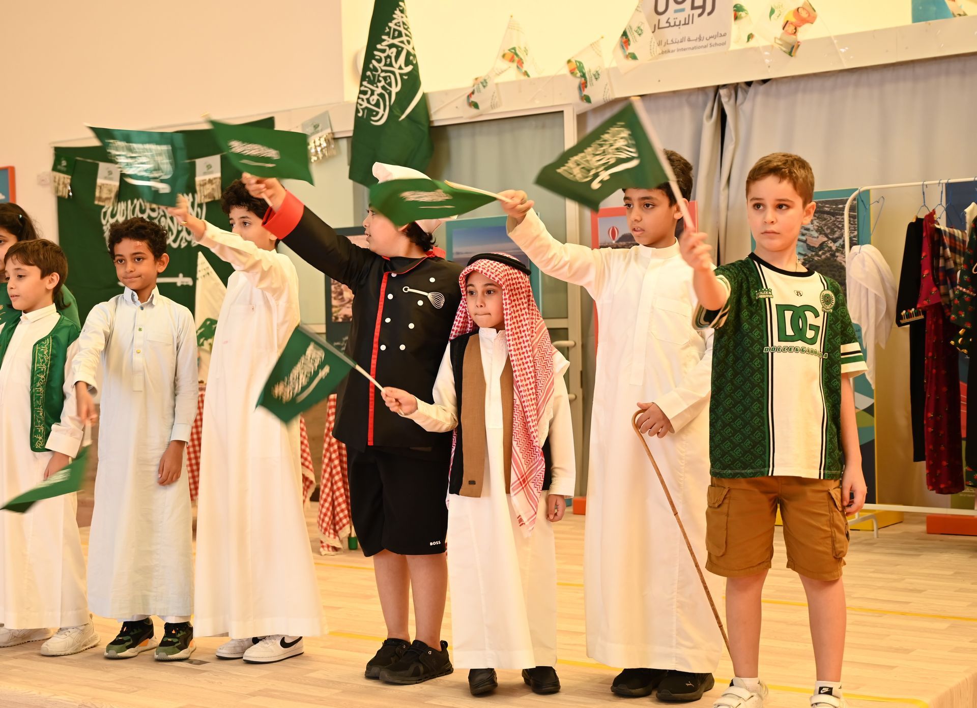 A group of children are standing on a stage holding Saudi flags.