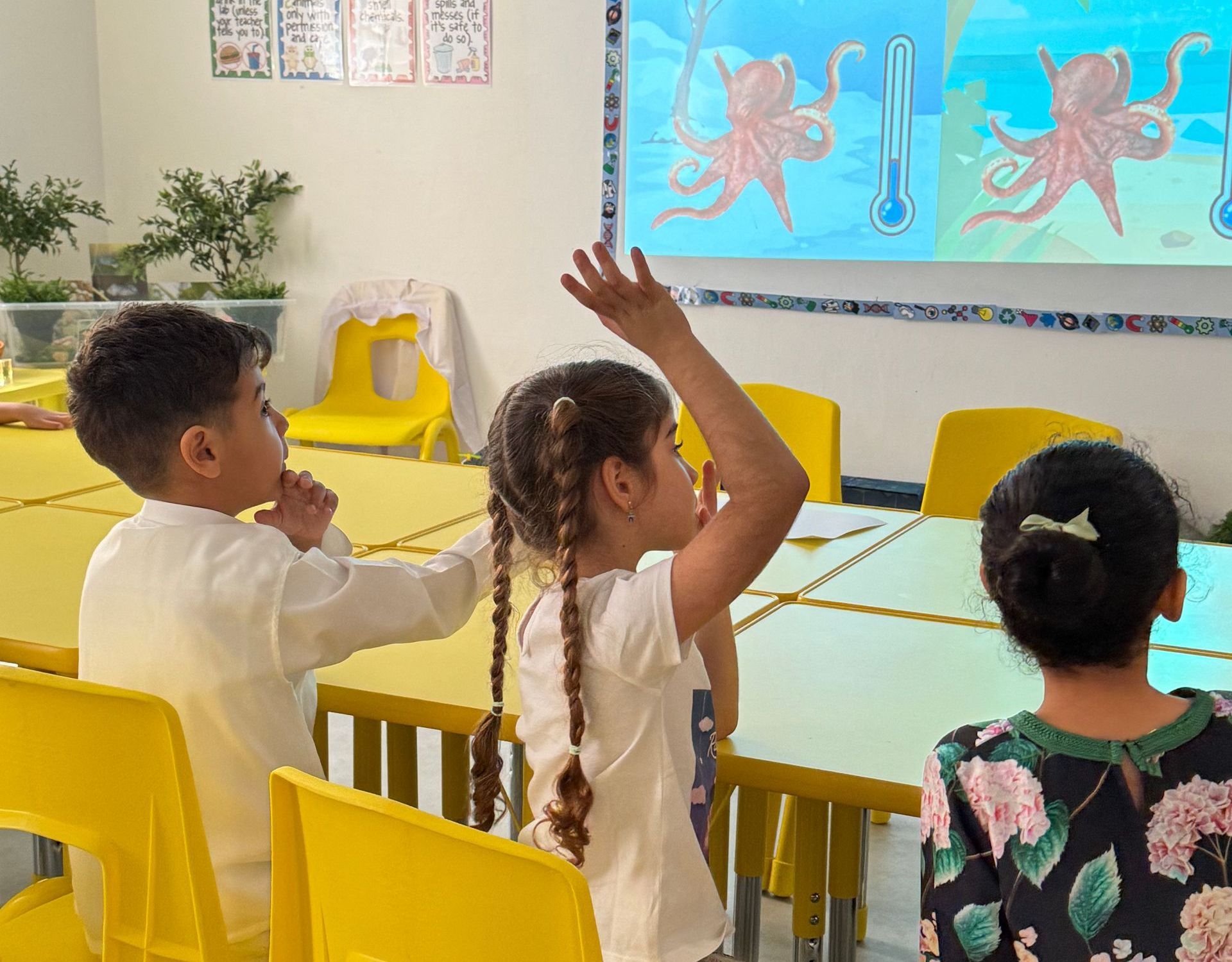 A group of children are sitting at tables in a classroom raising their hands to answer a question.