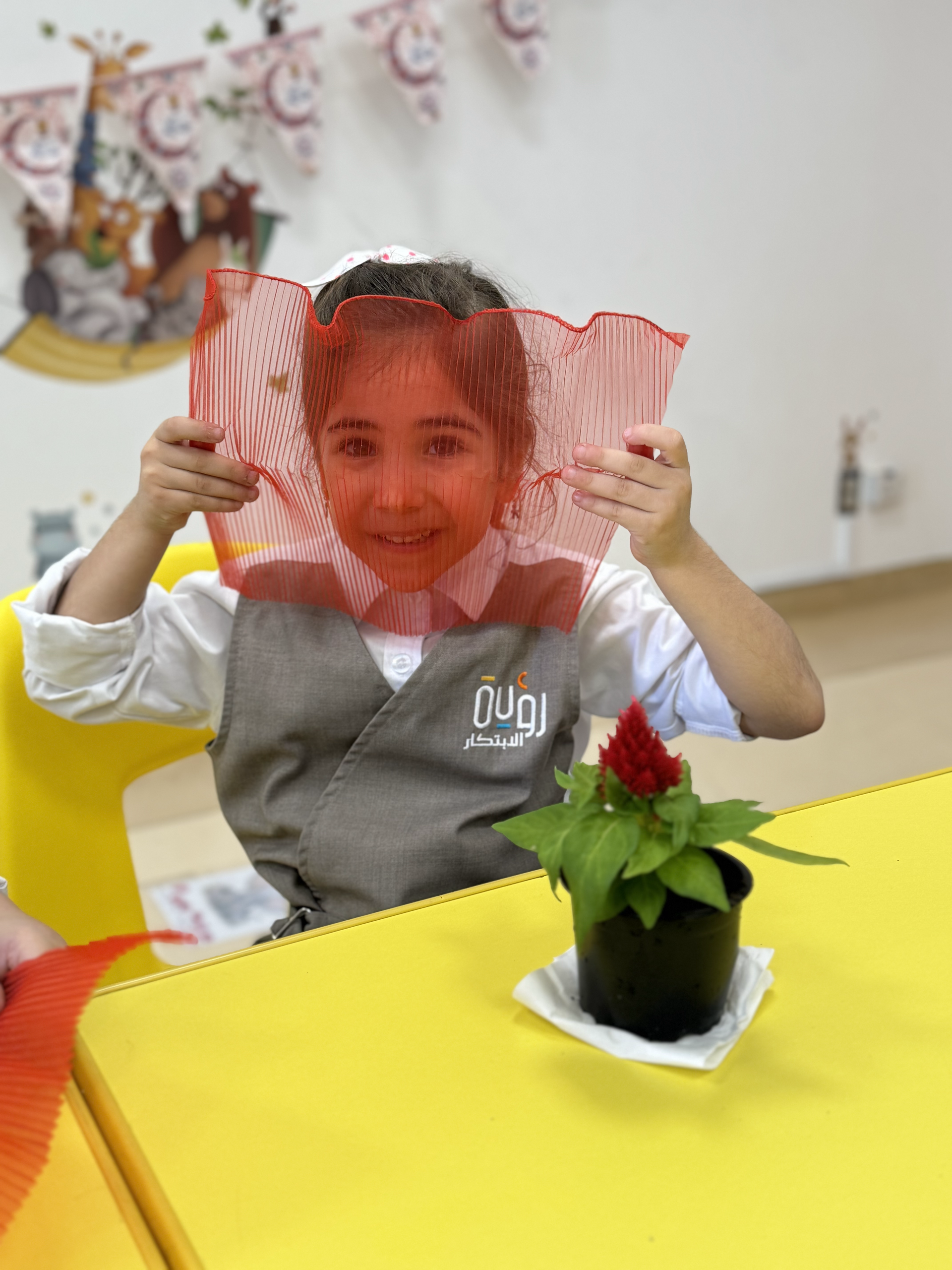 A little girl is sitting at a table with a potted plant in front of her face.