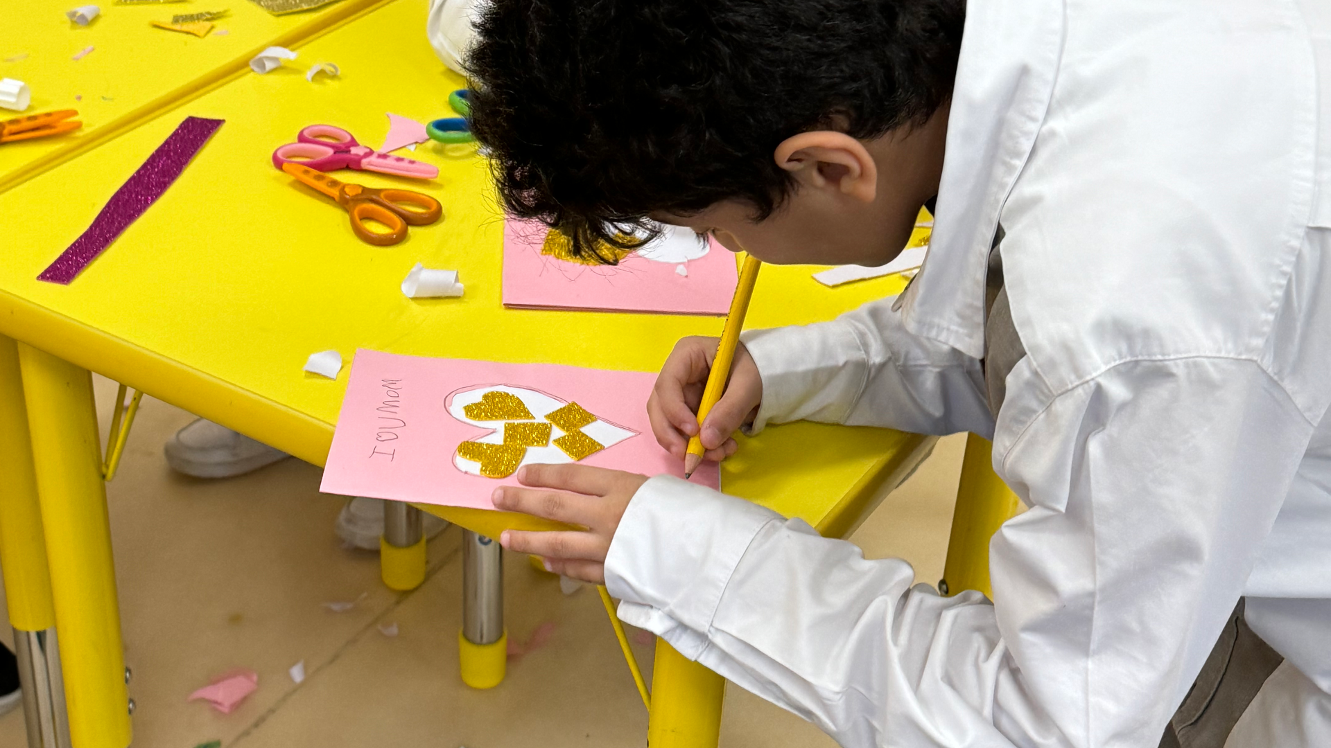 A young boy is sitting at a yellow table drawing on a piece of paper.