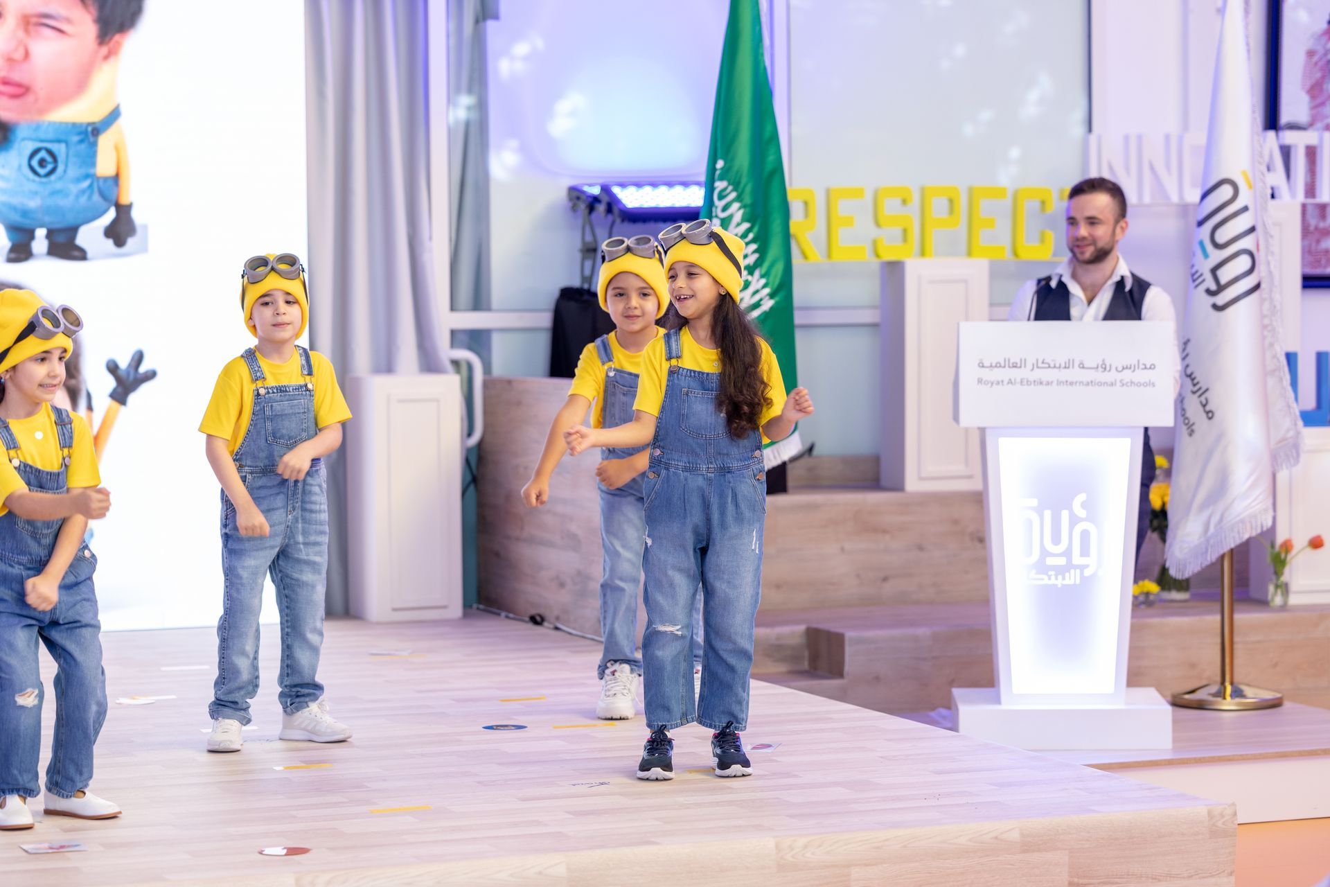 A group of children dressed as minions are dancing on a stage.