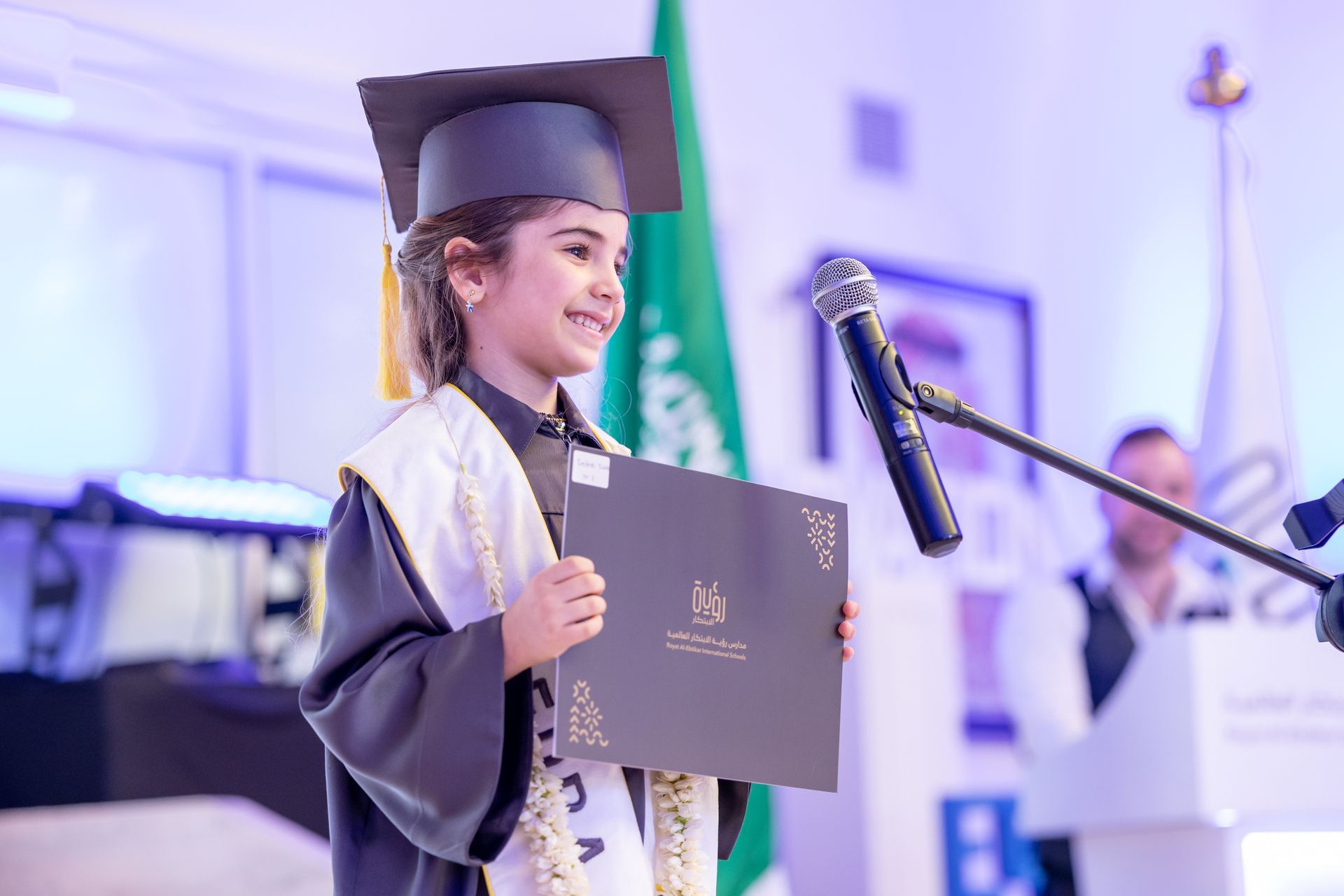 A young girl in a graduation cap and gown is holding a diploma in front of a microphone.