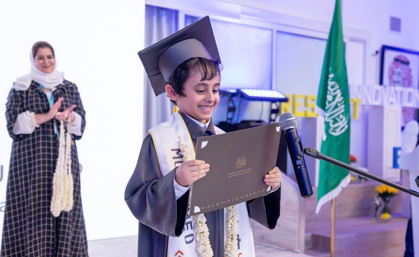 A young boy in a graduation cap and gown is holding a diploma in front of a microphone.