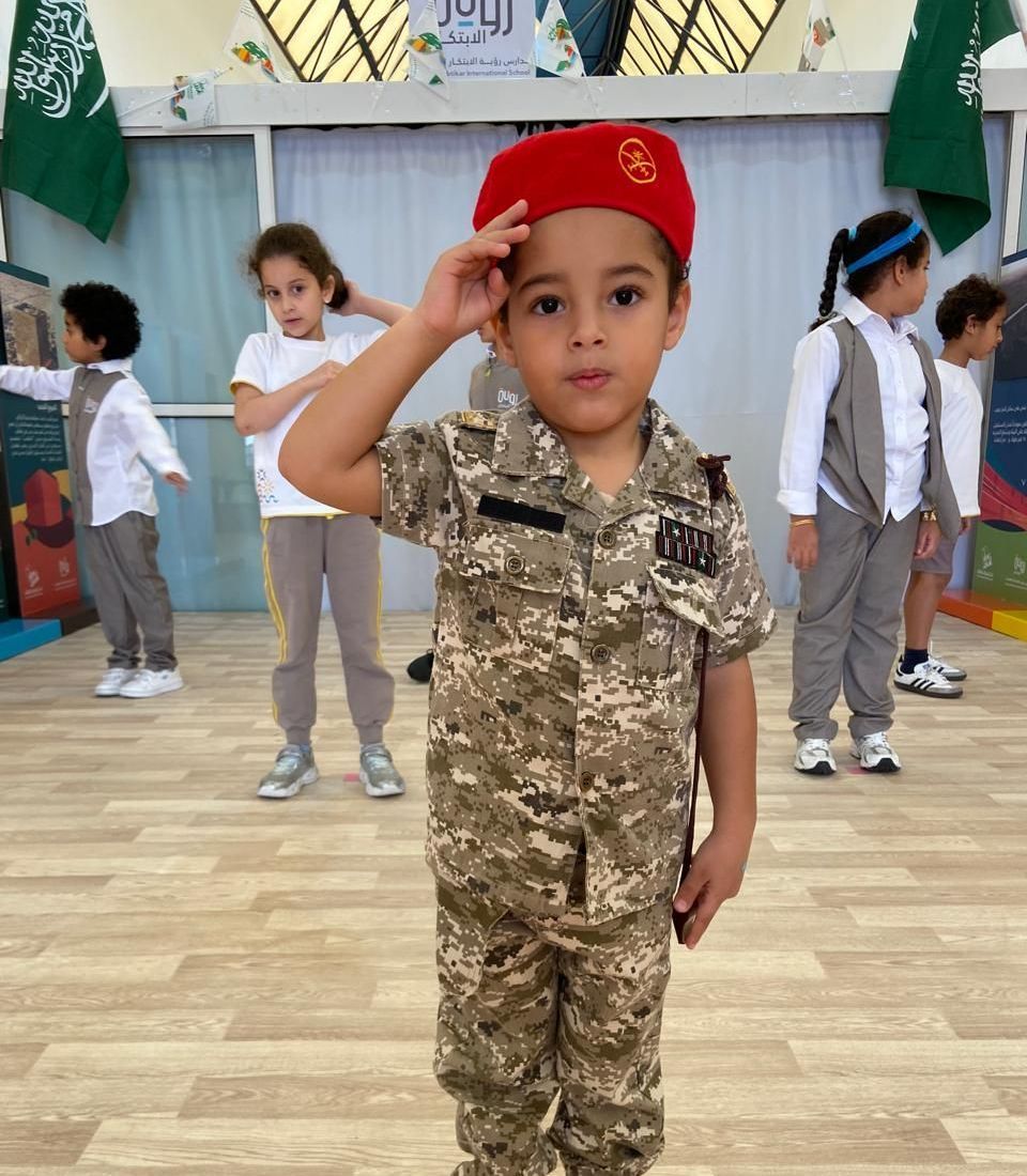 A young boy in the Saudi military uniform salutes in front of a group of children.