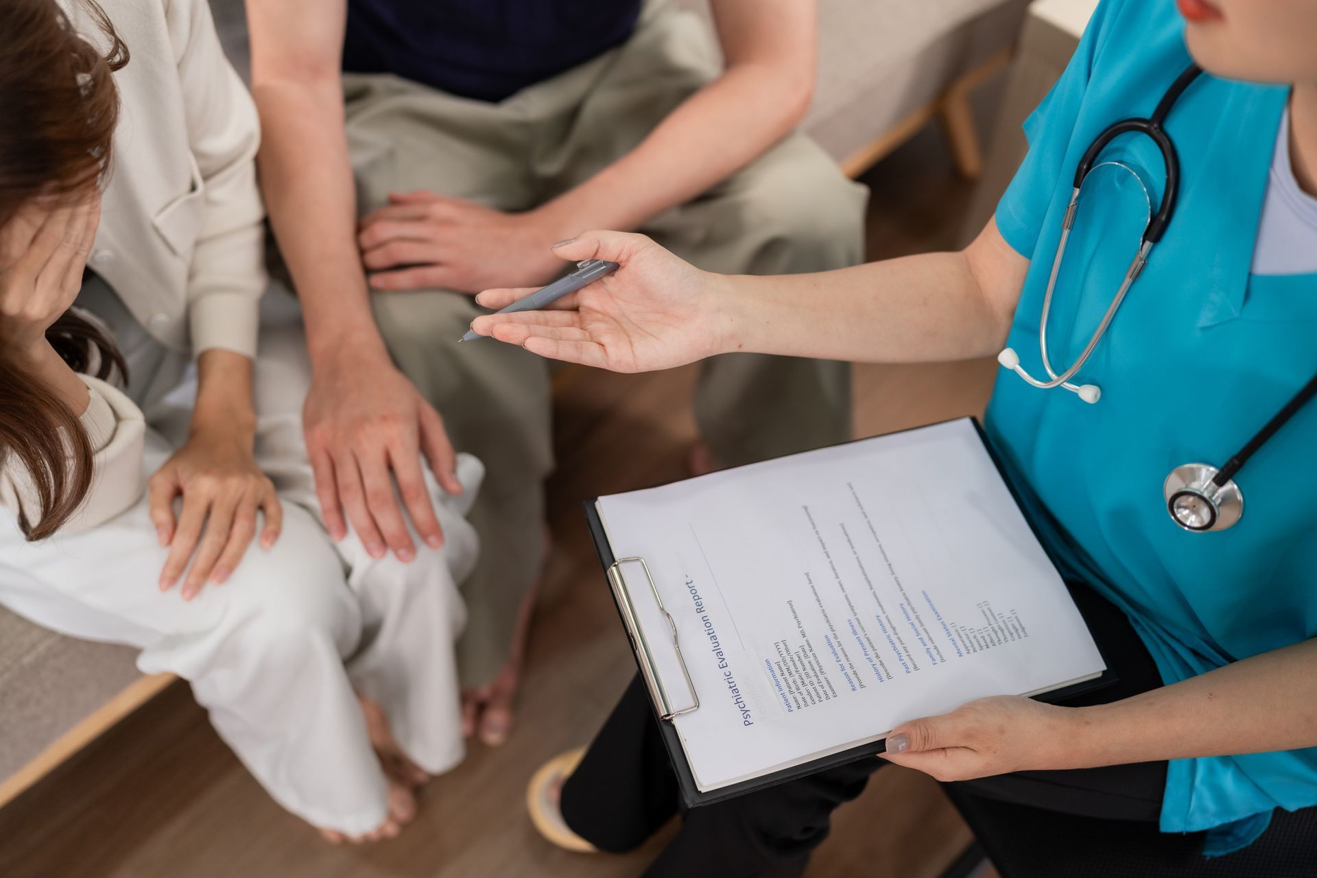 A healthcare professional in blue scrubs holding a clipboard talks to two individuals sitting on a couch.