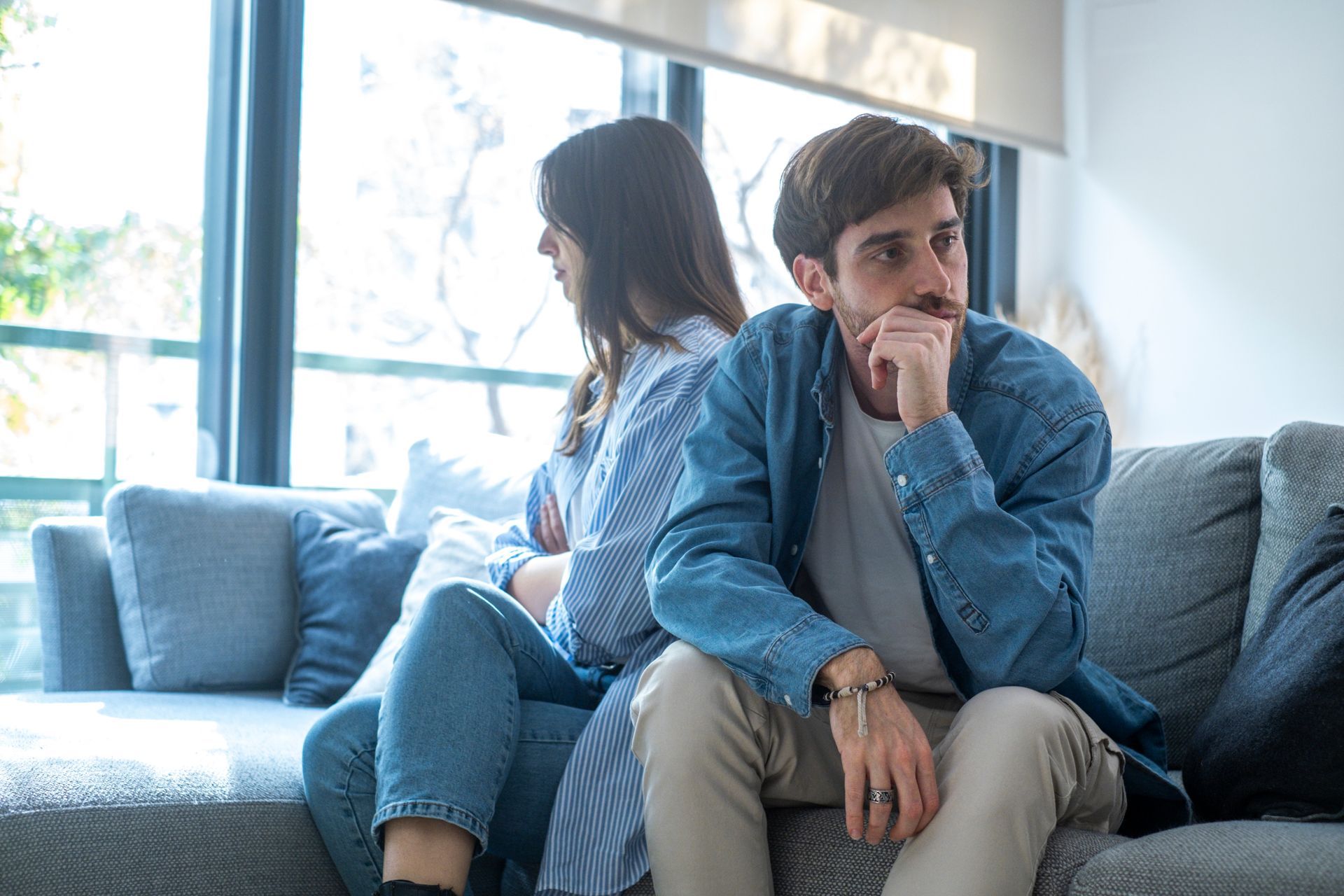 Couple sits back-to-back on couch, looking upset and distant after an argument at home in silence.