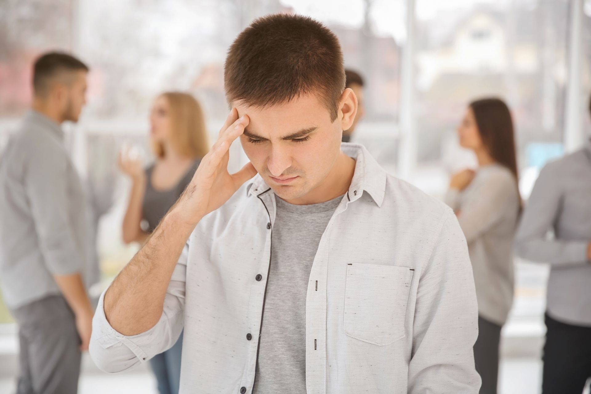 Young man holds his head, looking stressed while people talk blurred in background nearby scene